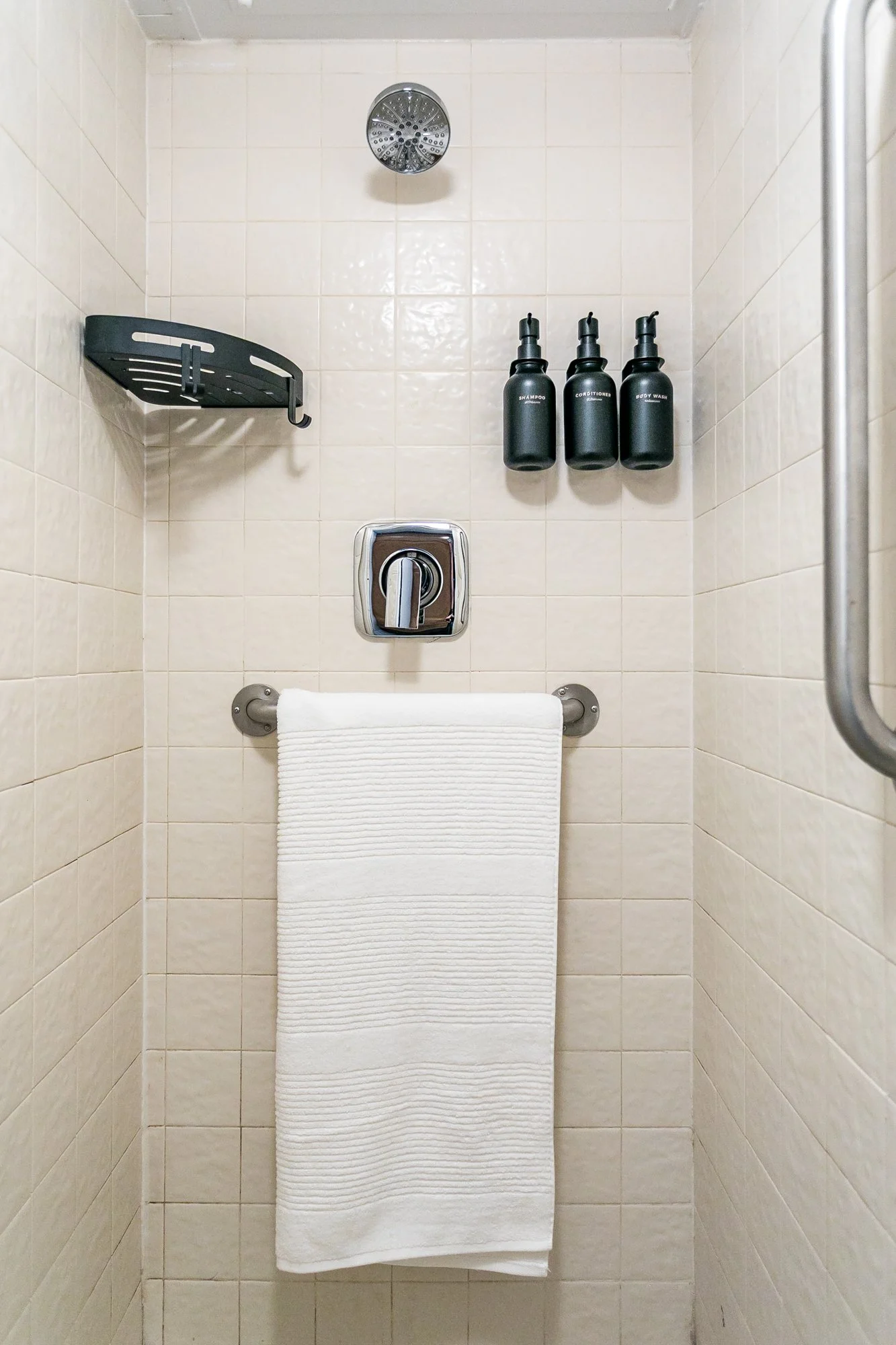 A shower stall with tiled walls, a showerhead, a shelf with hooks, three bottles labeled shampoo, conditioner, and body wash, a towel and a grab bar on the right side.