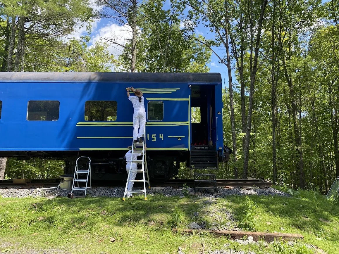 Two people painting yellow stripes on a blue train car beside a wooded area. One person is on a ladder, and the other is holding the ladder from below.
