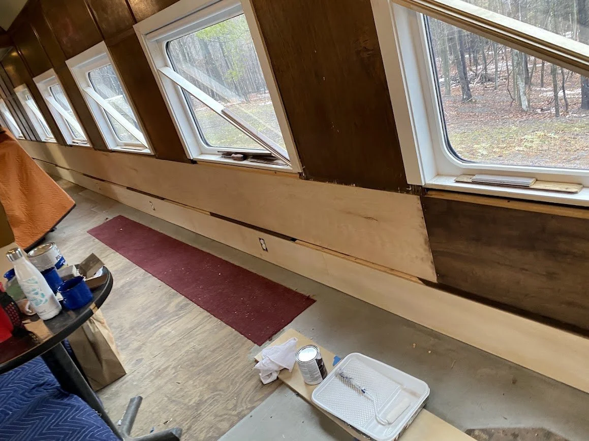 Interior of a room of a train under renovation with multiple windows, a red rug, painting supplies on the floor, and wood paneling.