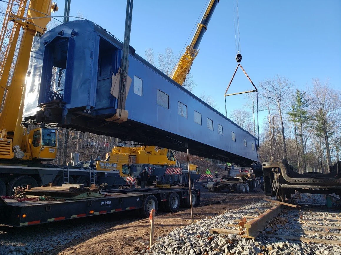 A large train car is being lifted by a yellow crane onto railway tracks at a construction site with workers present.