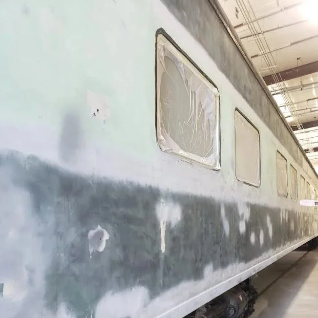 Close-up of a train car with a patched exterior, showing several windows, some covered or taped, in an indoor train maintenance area before it is repainted.