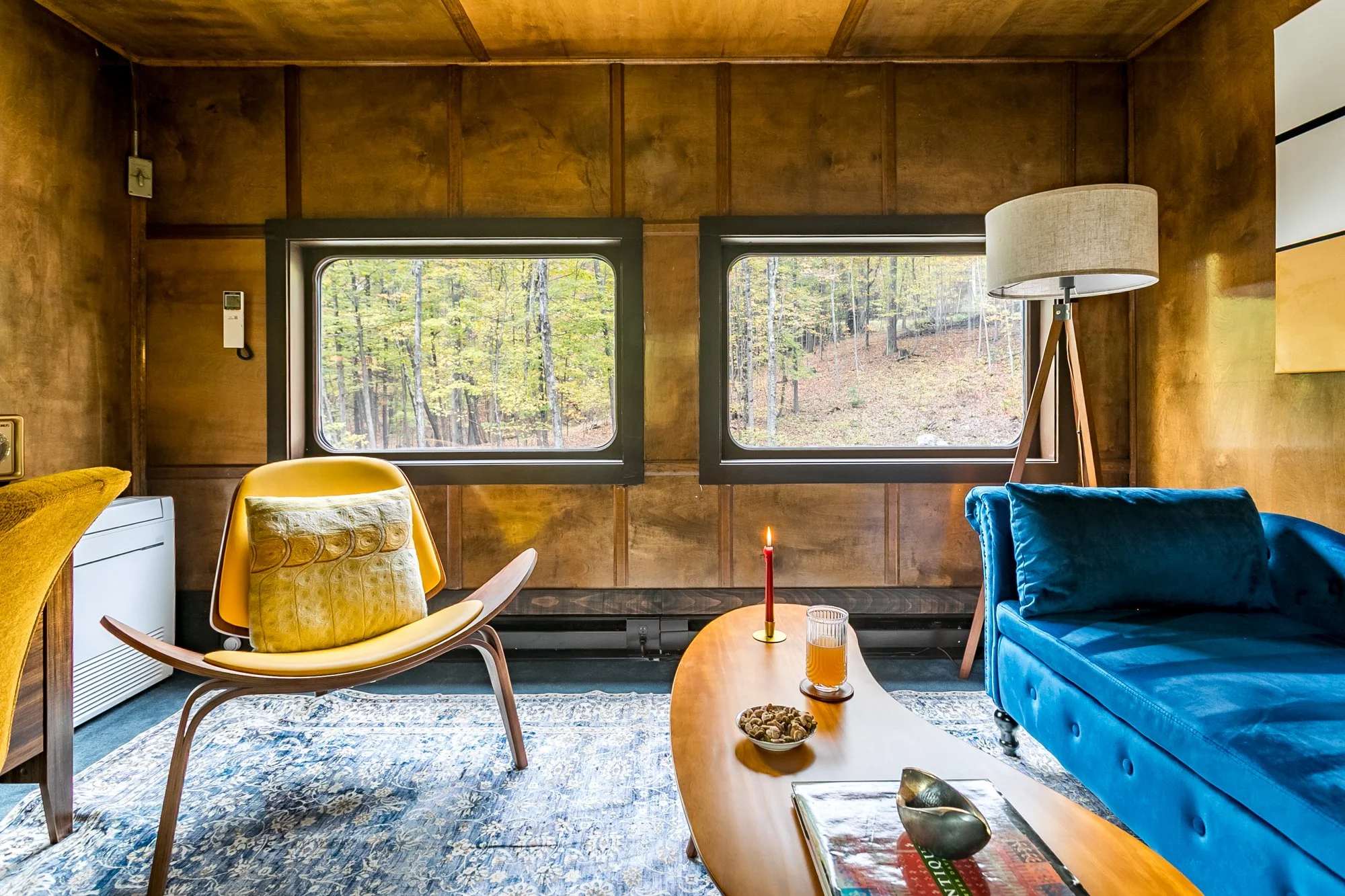 Wood-paneled study with two large windows showing a forest outside. The room has a yellow lounge chair and a blue velvet chaise sofa, a wooden coffee table and a tall floor lamp.