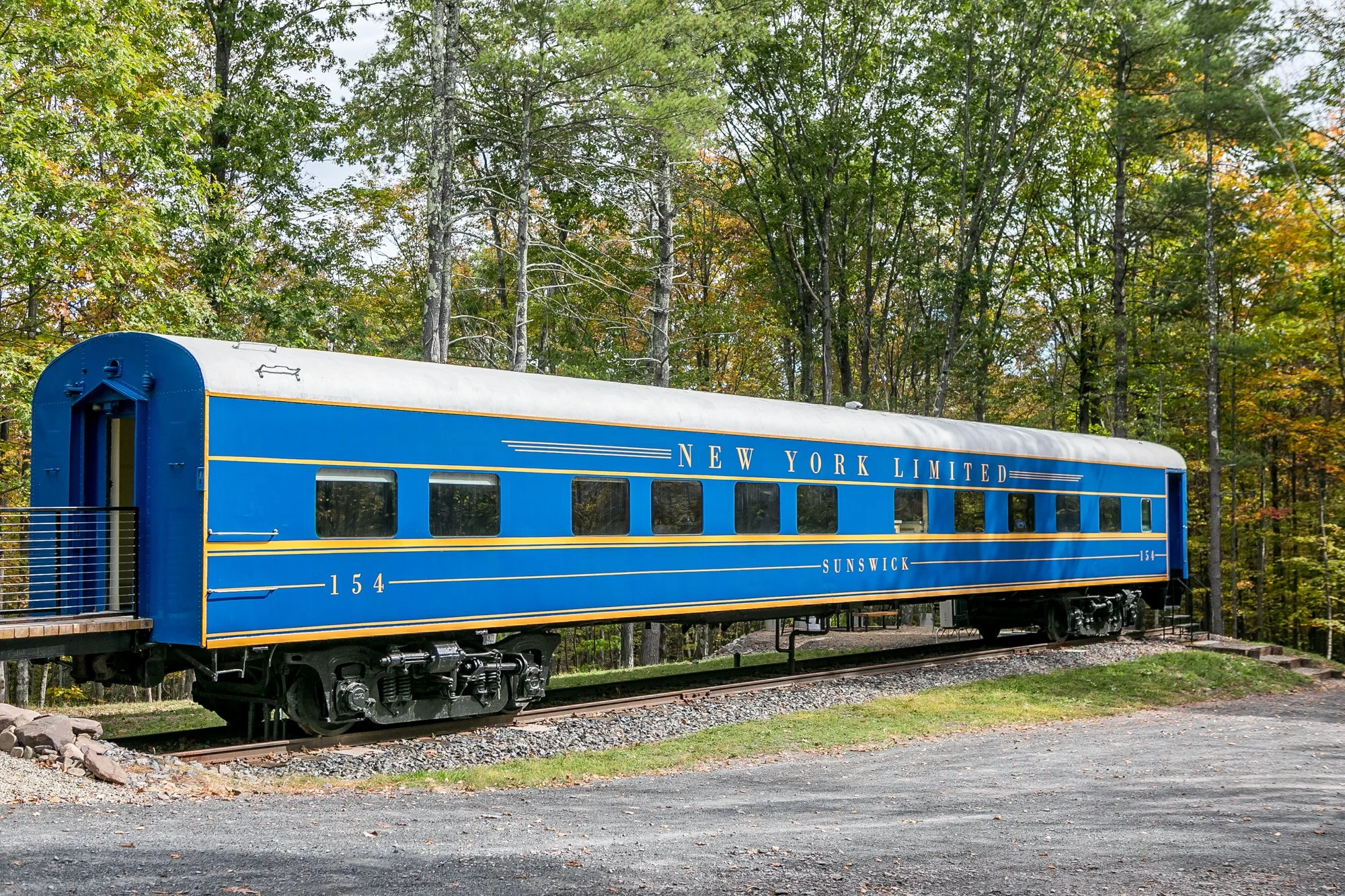 A vintage blue train car with the words 'New York Limited Sunswick' on its side, sitting on a railway track surrounded by trees with autumn foliage.
