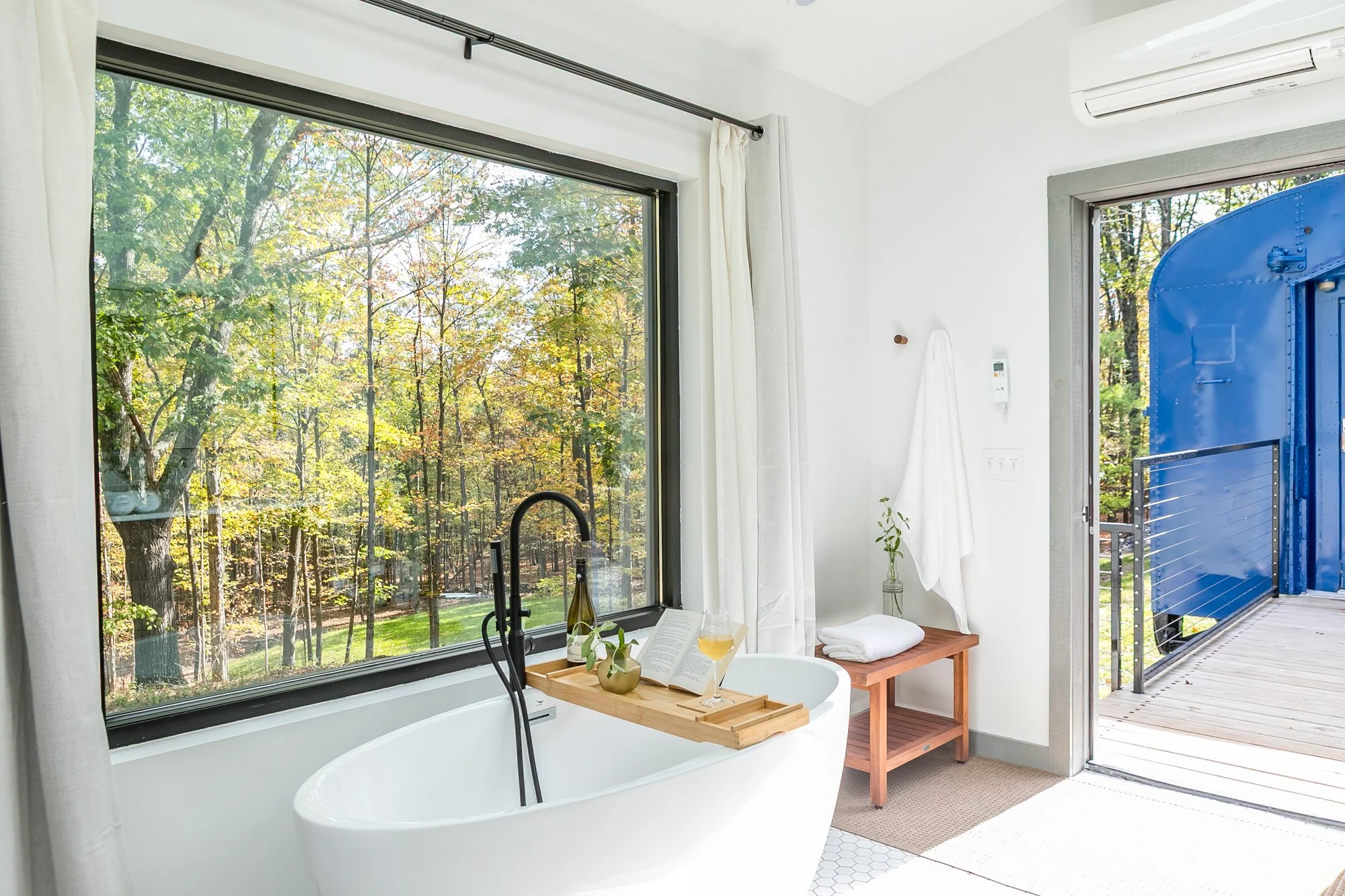 A bathroom with a large window showing a wooded scene, a freestanding bathtub with a wooden tray holding a book, glass of wine, and a bottle, a small wooden bench with towels, and an open door leading to a deck to the train with trees outside.