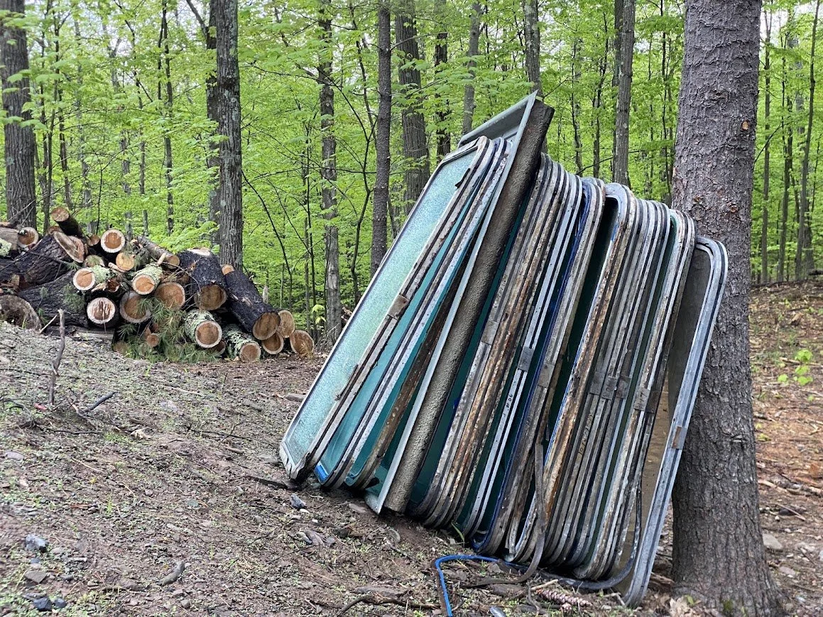 A stack of old, rusty windows leaning against a tree in a forested area with cut logs in the background.