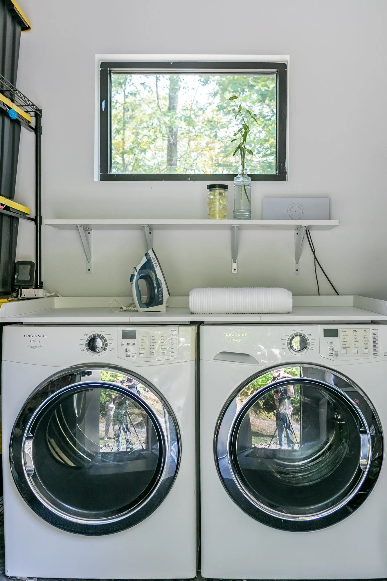 Front view of a laundry area with a front-loading washing machine and dryer side by side, a shelf above. A window above displays an outdoor scene with trees.