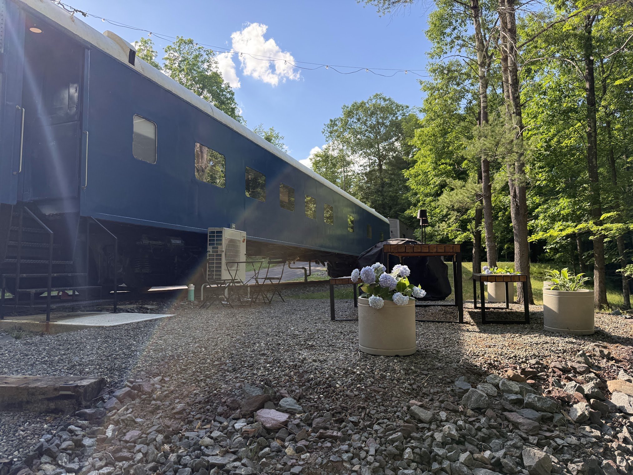 Outdoor scene with trees, a large blue train car, potted plants with white flowers, and gravel ground under a clear blue sky.