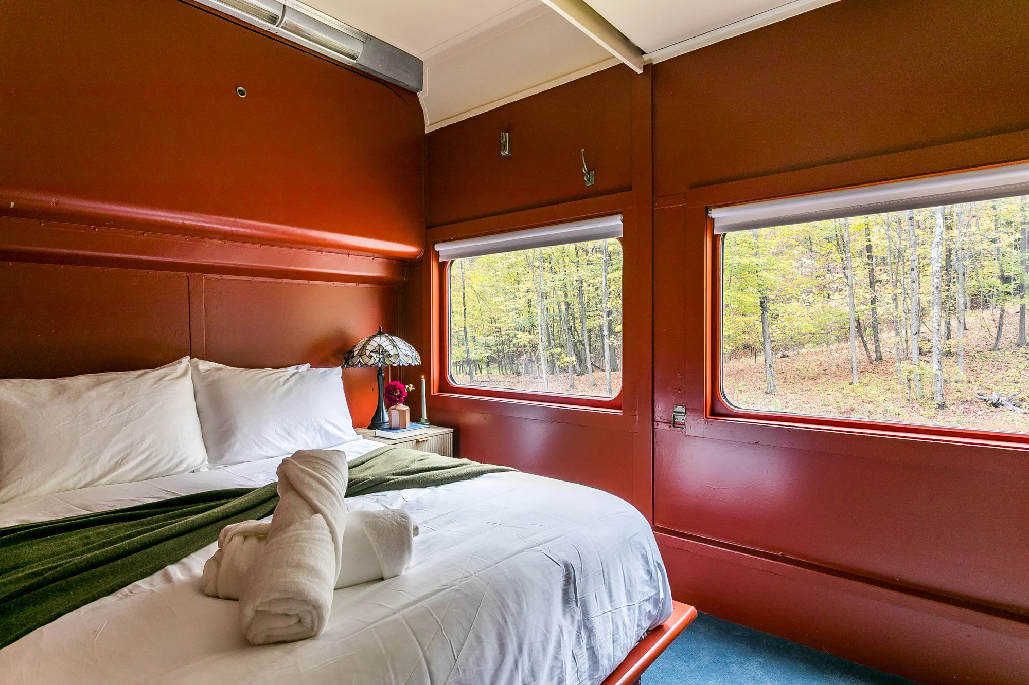A cozy bedroom with a bed covered in white sheets and green blanket, a folded towel shaped like a swan, a bedside table with a colorful stained glass lamp, and large windows showcasing a forest with trees and greenery outside.