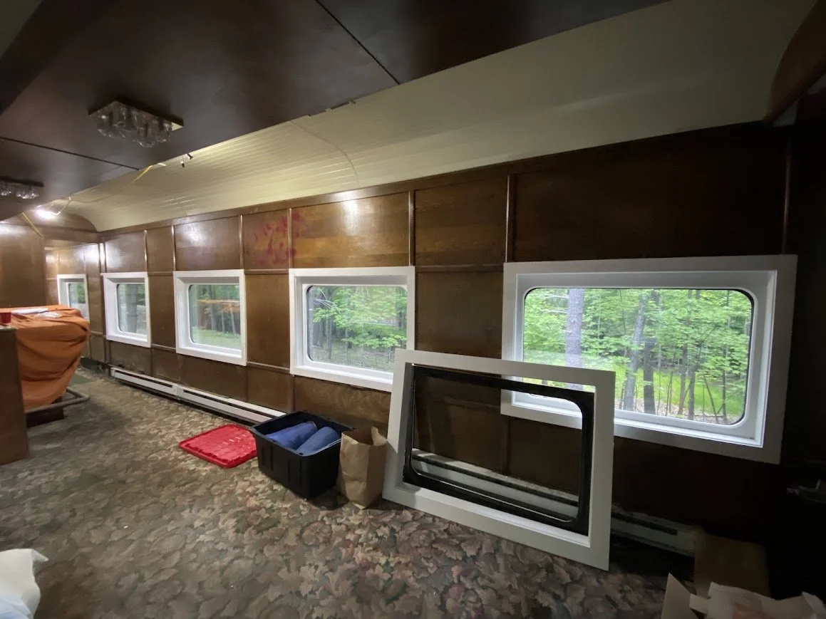Inside a room with a row of windows revealing a green forest outside, with construction or renovation materials and boxes on the floor.