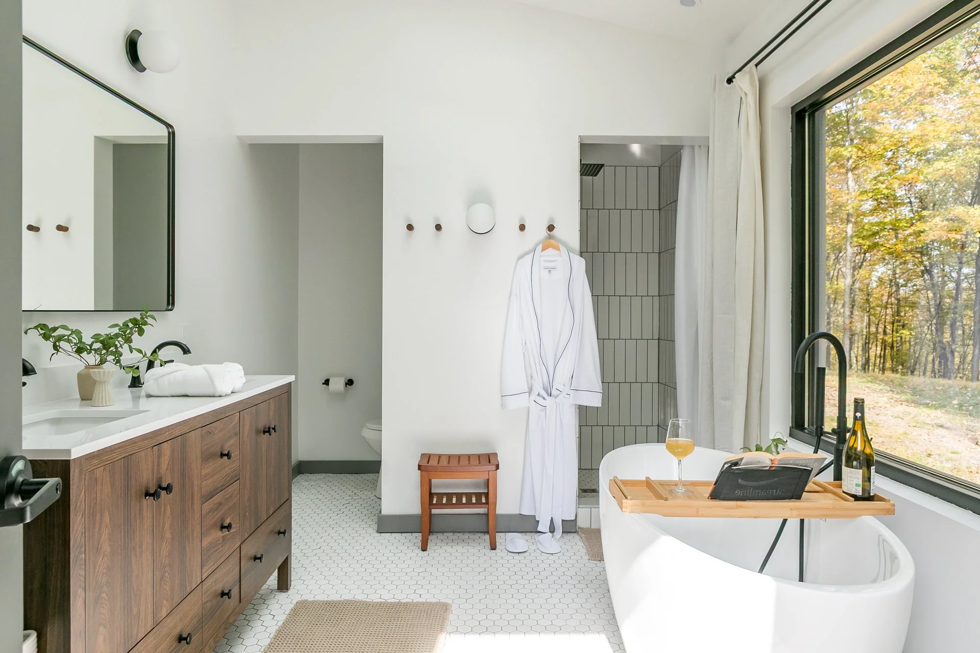A modern bathroom with a picture window, a large soaking bathtub, walk-in shower with rain showerhead and white robes.