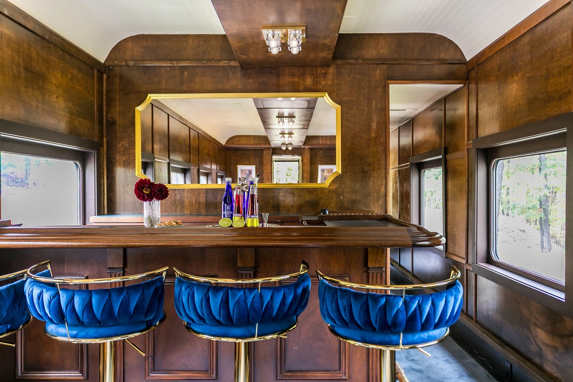Interior of a vintage train car with wooden paneling, a mirror on the wall, and blue velvet bar stools along a vintage wooden bar.