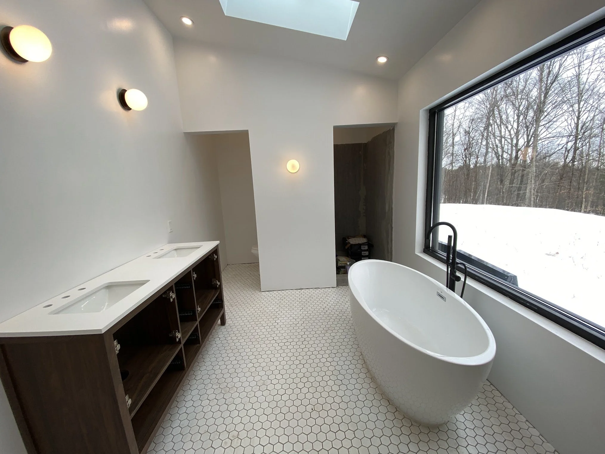 A modern bathroom with a freestanding bathtub by a large window showing snow-covered trees outside. The bathroom has a white wall with built-in lighting, a skylight, and a vanity with under-mount sinks and dark cabinetry.