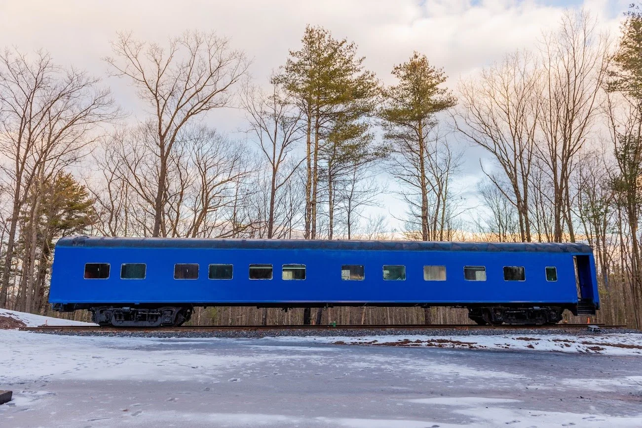A blue train car on train tracks in a winter landscape with snow and a background of leafless trees and a cloudy sky.