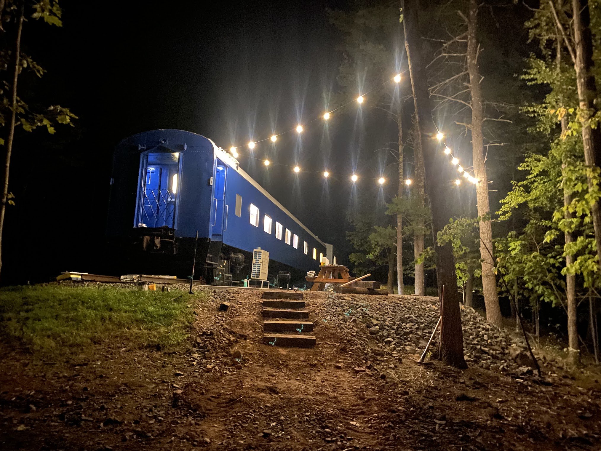 Nighttime scene of a blue train carriage on a track, illuminated string lights overhead, surrounded by trees, with stairs leading toward the train. 