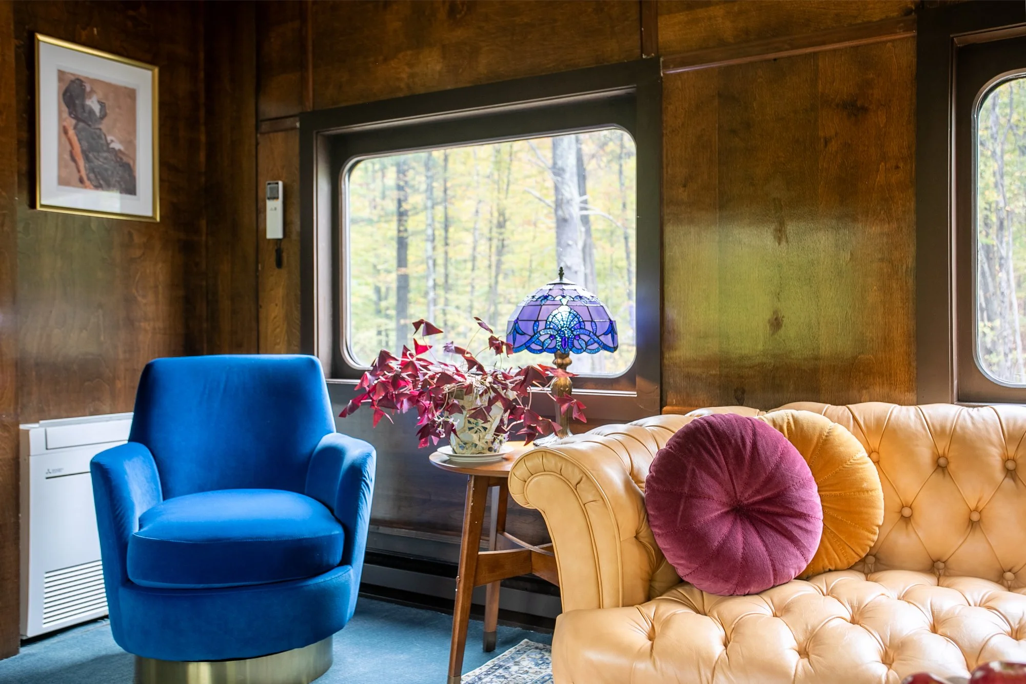 A vintage-style living room with wood-paneled walls, a large window showing trees outside, a blue armchair, a leather tufted sofa with pillows, a small wooden side table holding a stained glass lamp and a plant, and a framed picture on the wall.