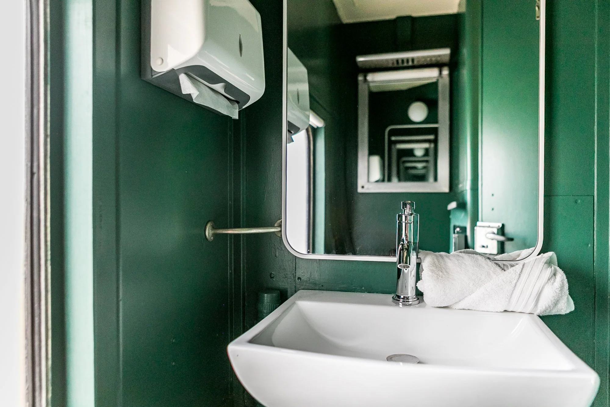 Green bathroom with a white sink, chrome faucet, mirror, and folded white towel on the sink. A soap dispenser is visible next to the sink.
