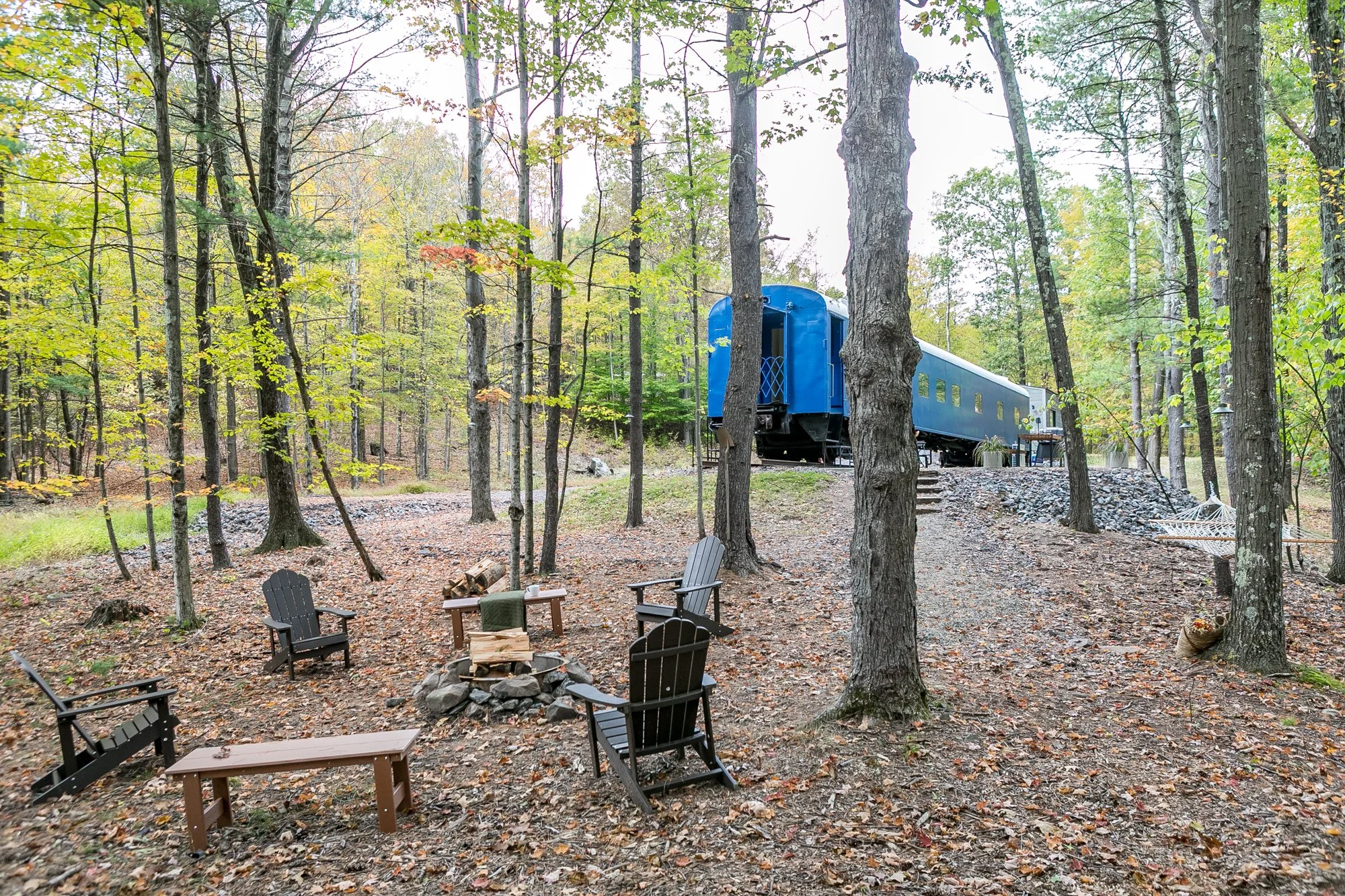 A wooded campsite with a blue train car as a shelter, surrounded by trees with green and yellow leaves, outdoor chairs around a fire pit, and some logs and a hammock.