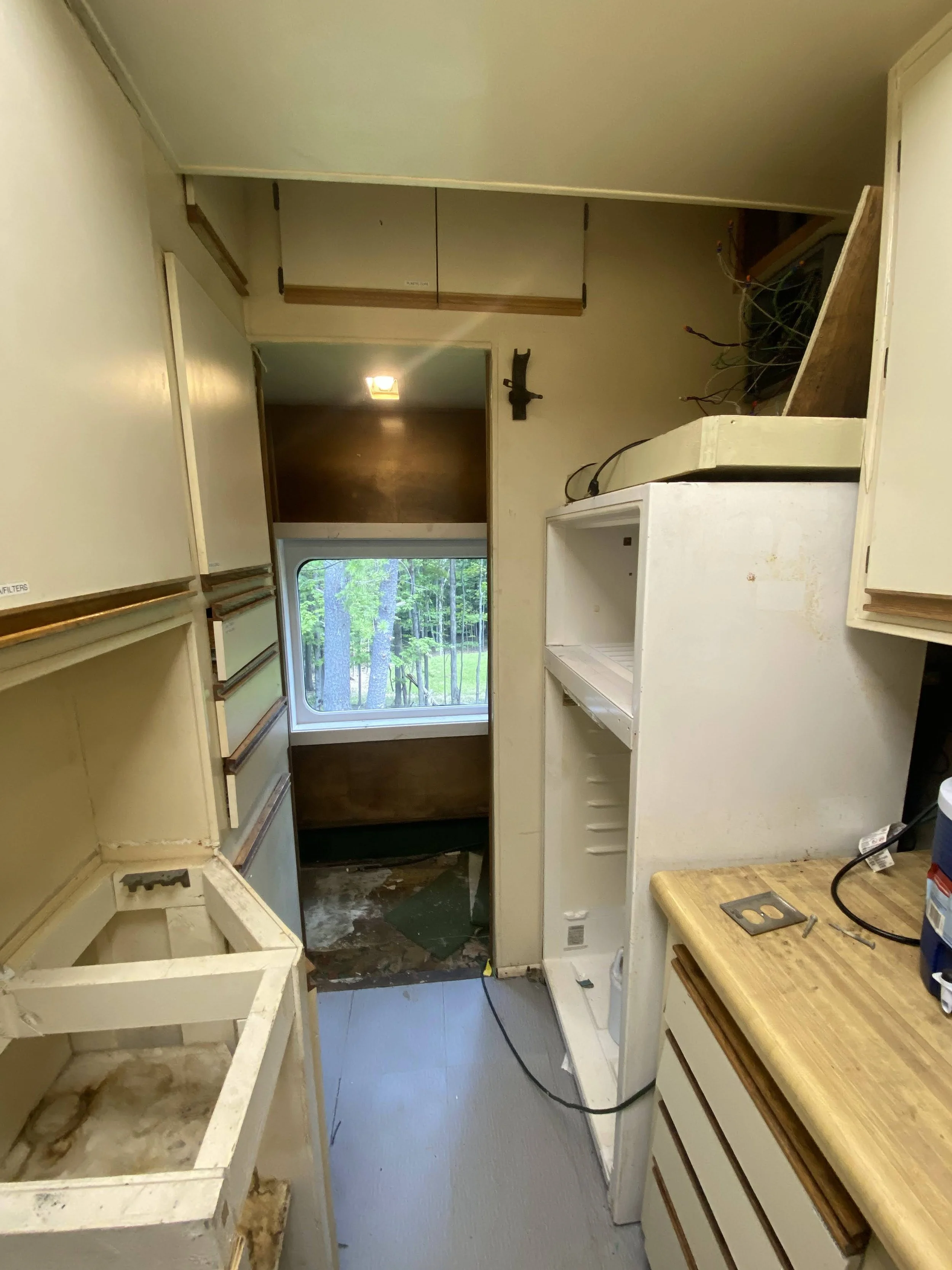 A kitchen under renovation with exposed wiring, empty cabinets, and a view outside through the window of trees and greenery.