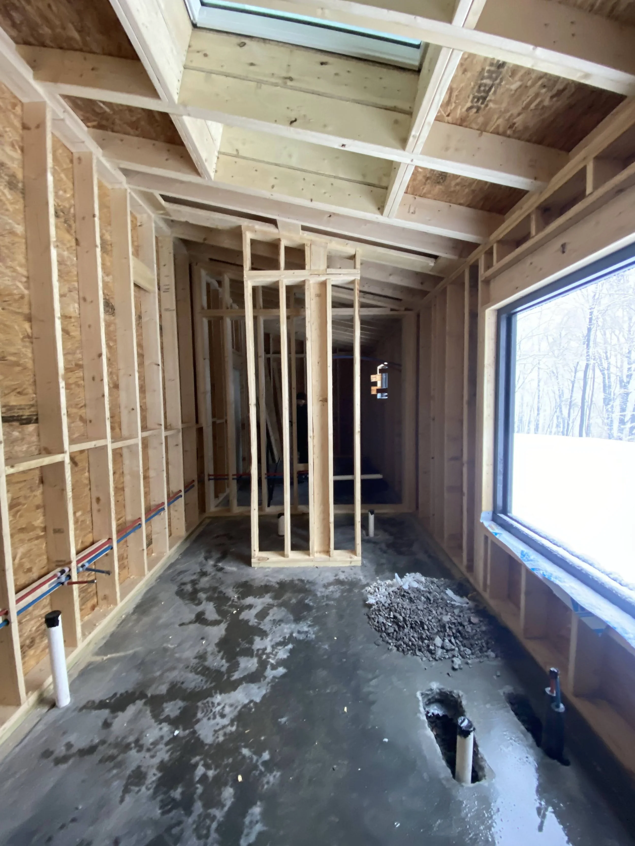 Interior view of a bath house under construction with wooden framing and a large window showing an outside winter scene.