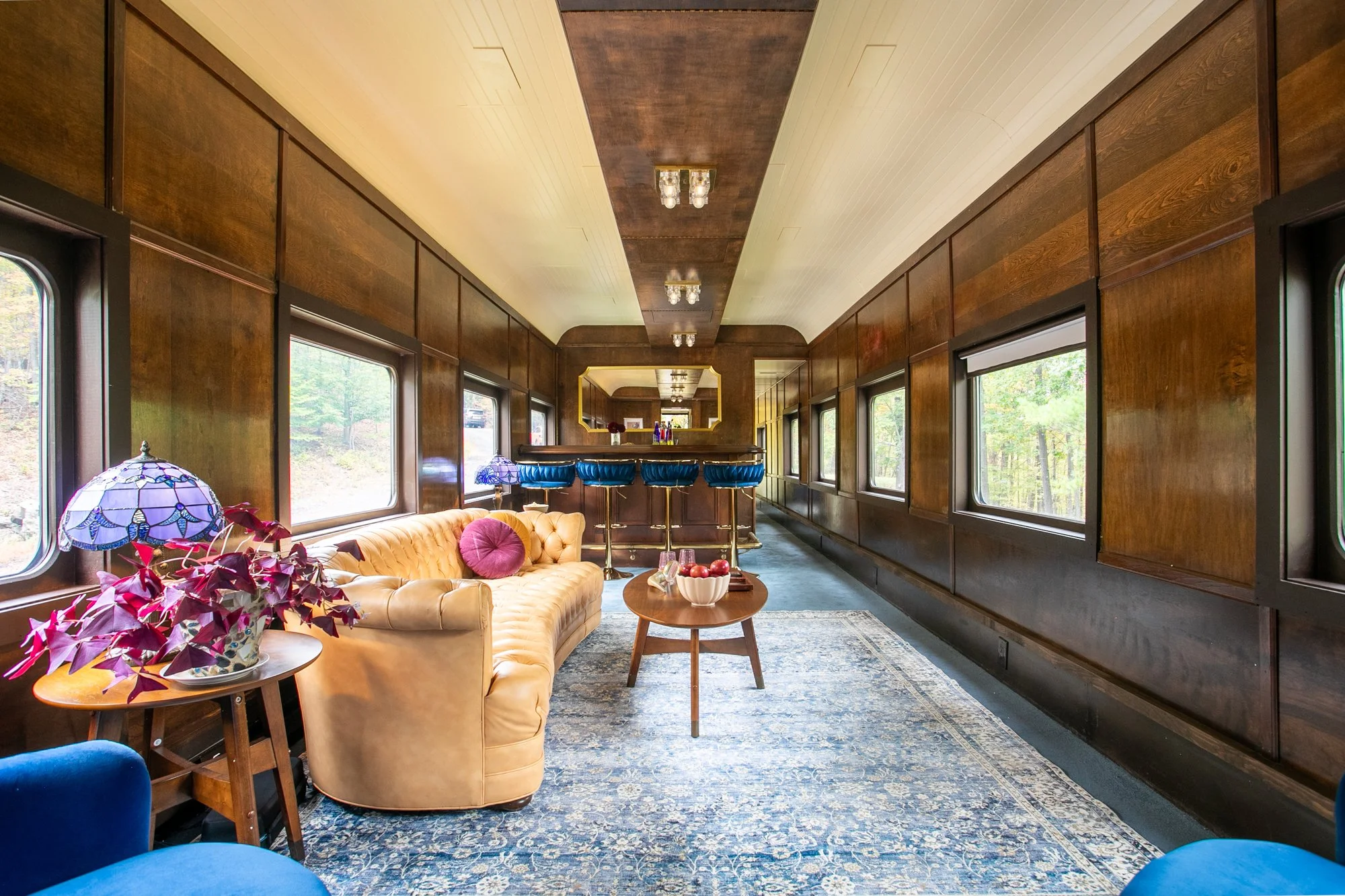 Lounge with wood-paneled walls, large windows, a custom leather tufted sofa with pink and blue pillows, a coffee table, blue club chairs. The room is decorated with vintage lamps and a mirror.