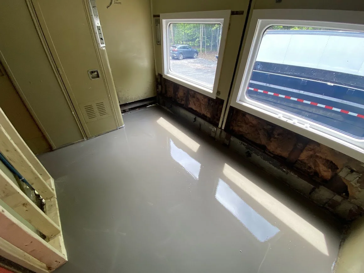 Room with a freshly poured concrete floor, studs for a wall, and large windows revealing an outdoor parking lot and trees.
