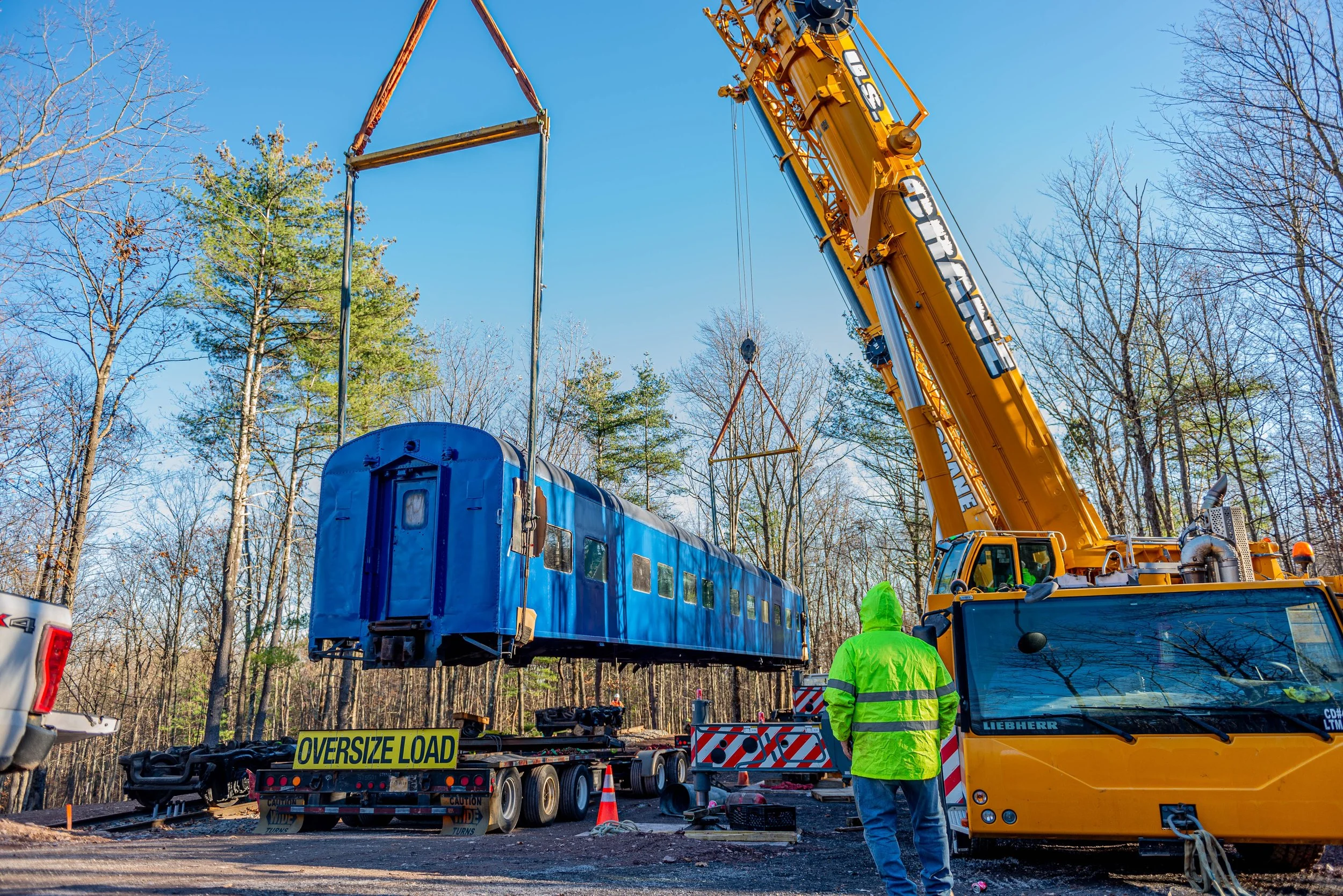 A construction site with a large yellow crane lifting a blue train car. Construction workers wearing neon green jackets and helmets are present, and various equipment and signs such as 'Oversize Load' can be seen amidst trees with no leaves and a clear blue sky.