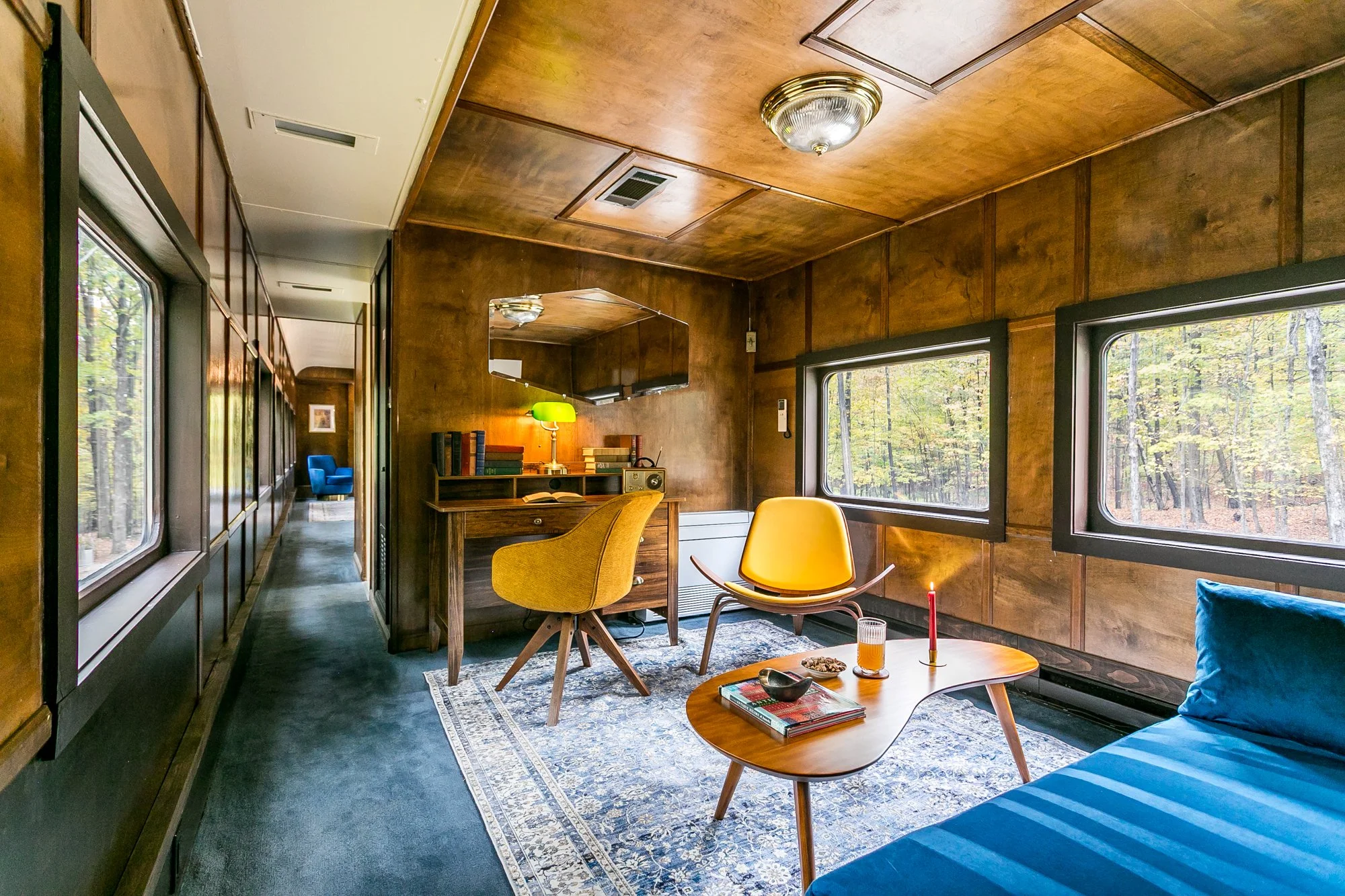 A cozy study with wood-paneled walls and large windows overlooking a forested outdoor scene. The room features a yellow chair and a round wooden table. There is a bookshelf, desk lamp, and a vintage radio. A blue sofa is partially visible.