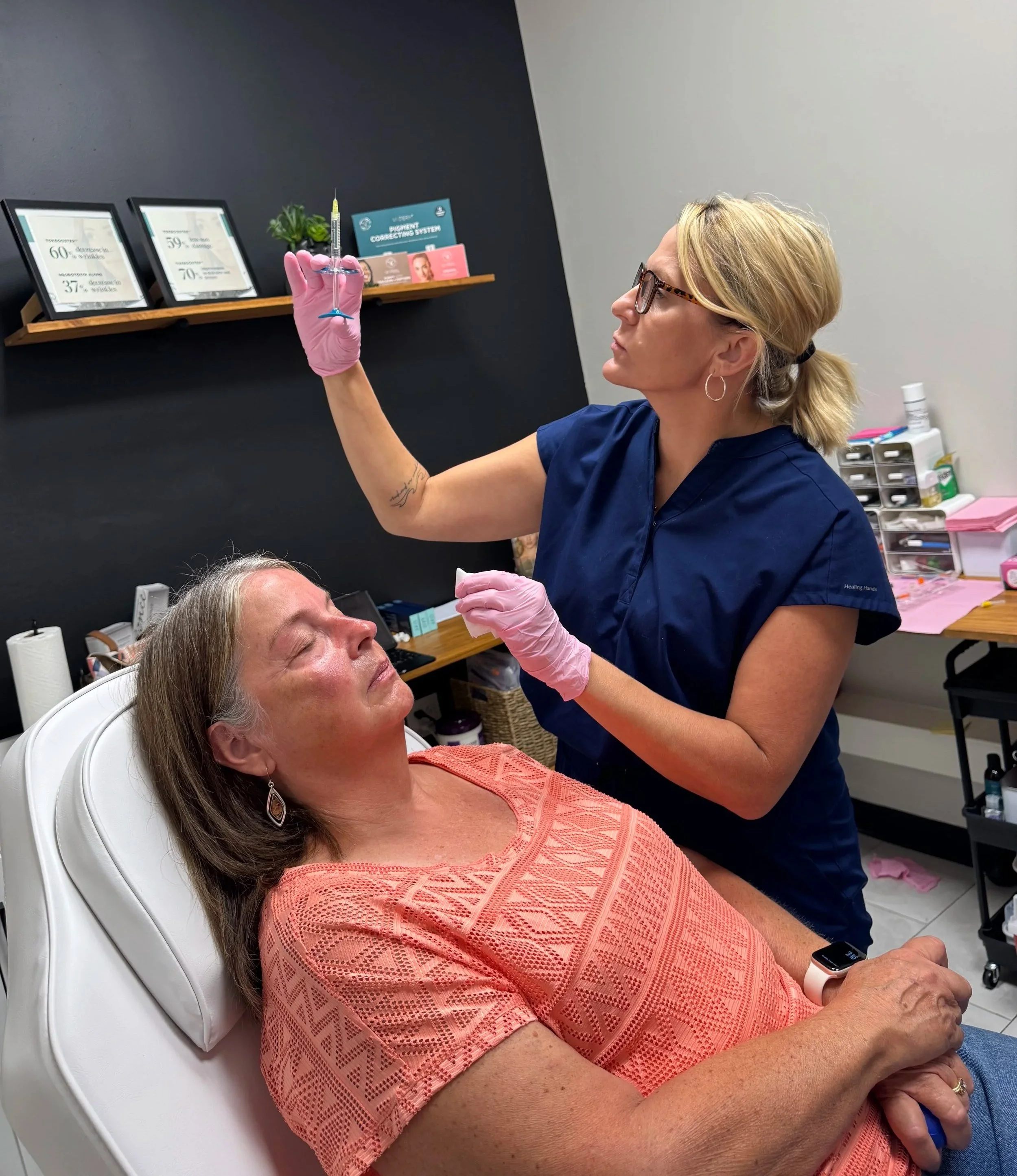 A woman with light brown hair and earrings is sitting in a medical office, holding a smartwatch, with her eyes closed. A healthcare worker wearing glasses and pink gloves is preparing a syringe, likely for an injection.
