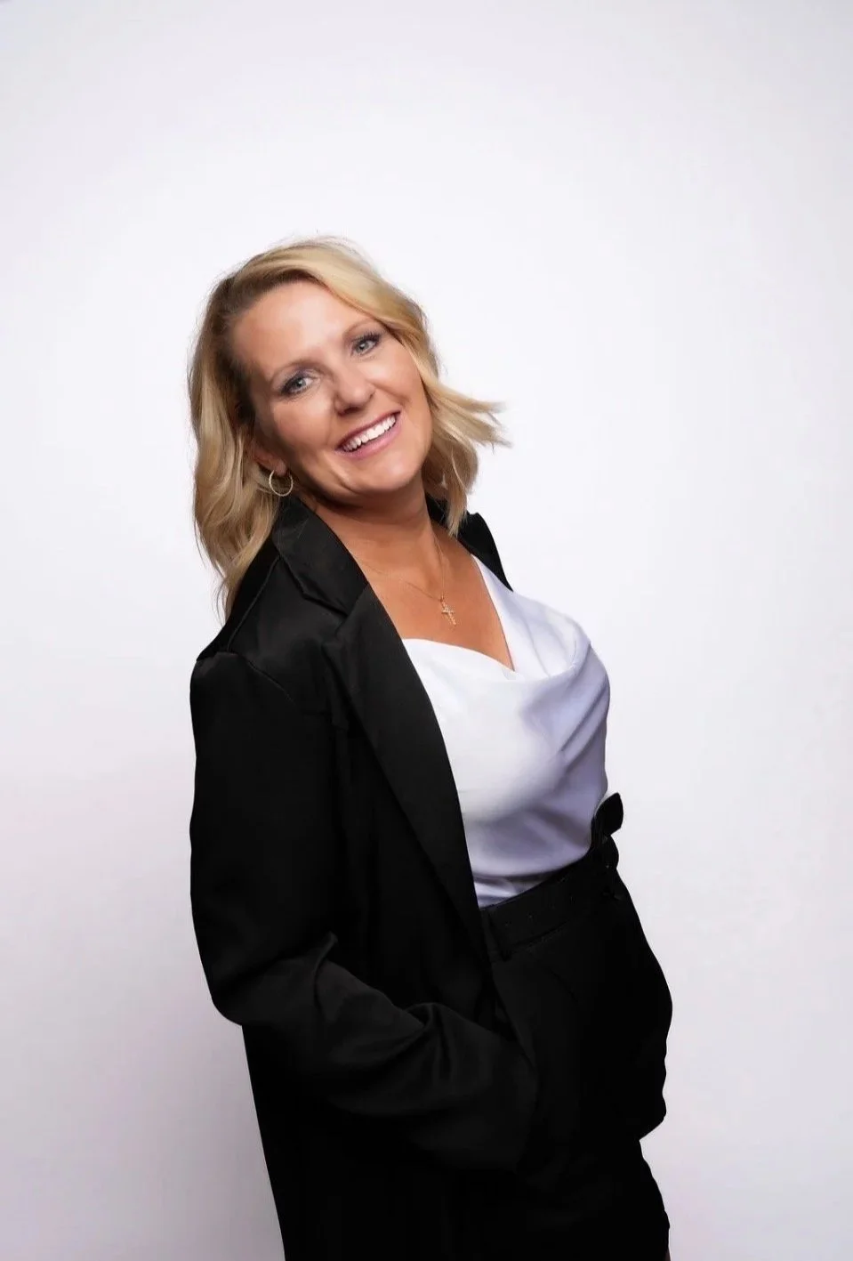 Smiling blonde woman with shoulder-length hair wearing a black blazer, white blouse, and black pants, standing against a plain white background.