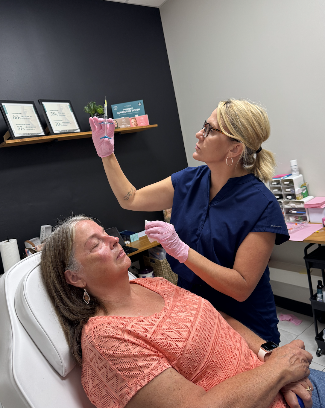 A medical professional in blue scrubs and pink gloves preparing a vaccination shot for a seated woman in an examination room.