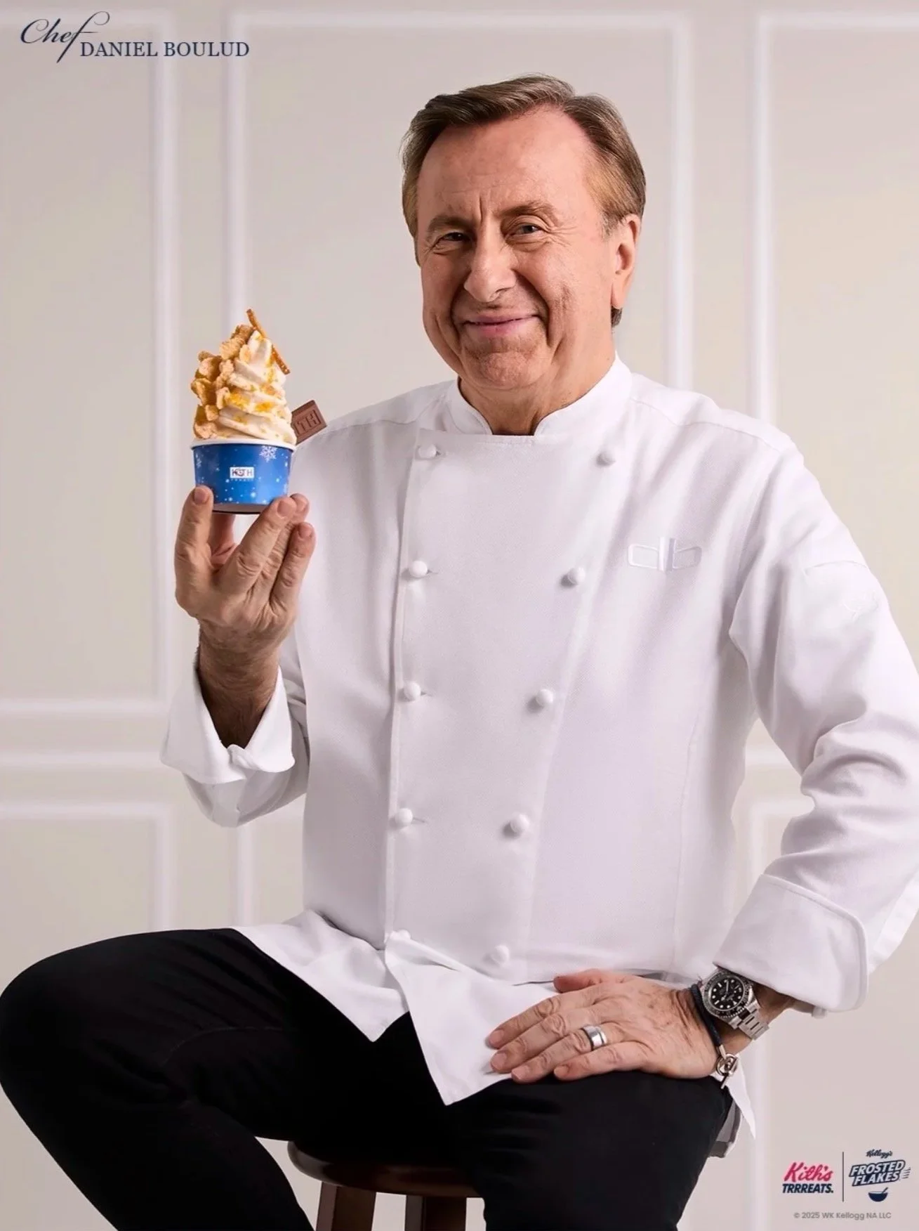 Chef Daniel Boulud in a white chef's coat holding a cup of frozen dessert topped with caramel and chocolate pieces, sitting on a stool in a white room.