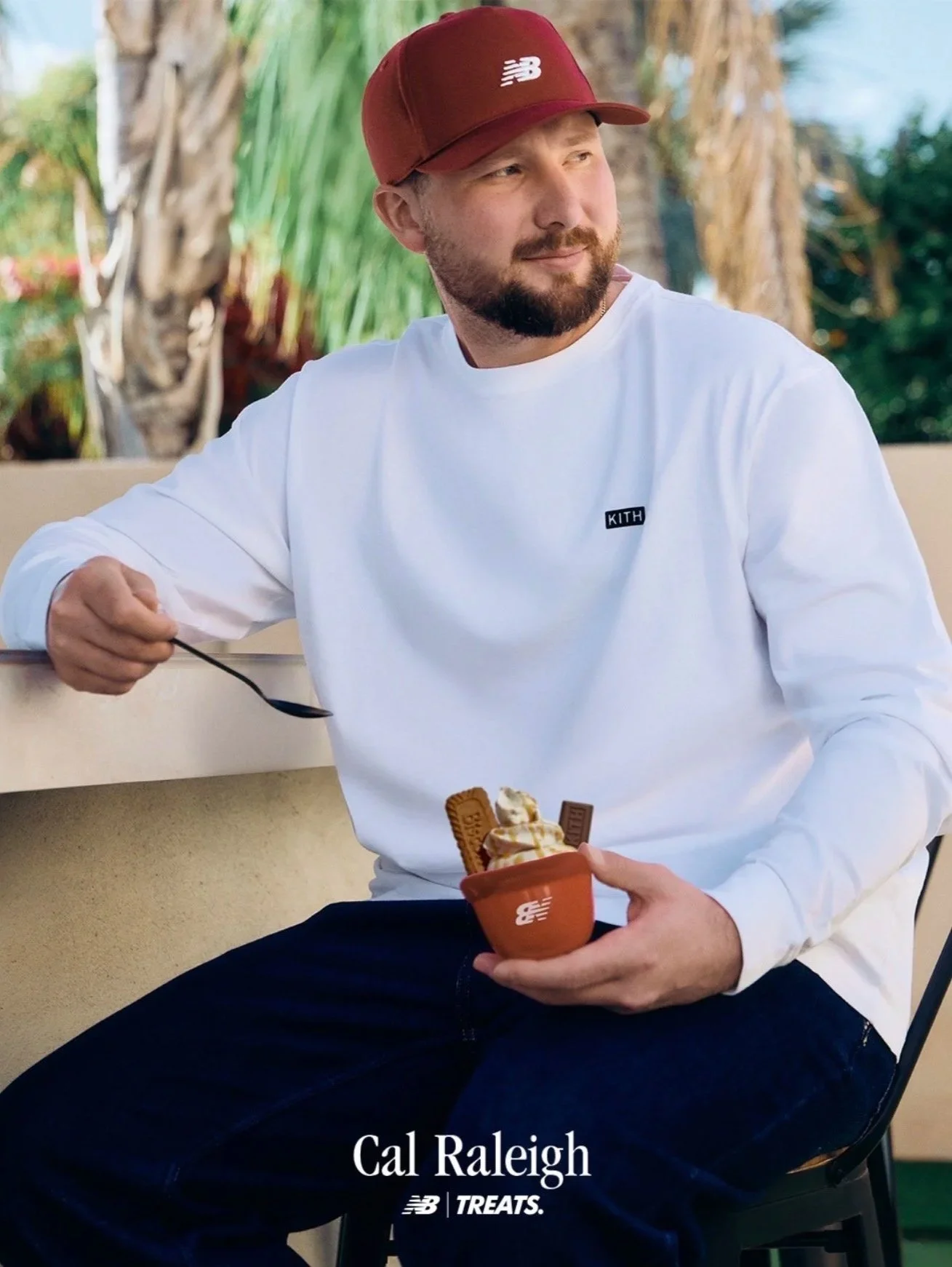 A man wearing a red New Balance cap and a white Kith sweatshirt sitting outdoors with palm trees in the background, holding a cup of ice cream with wafers.