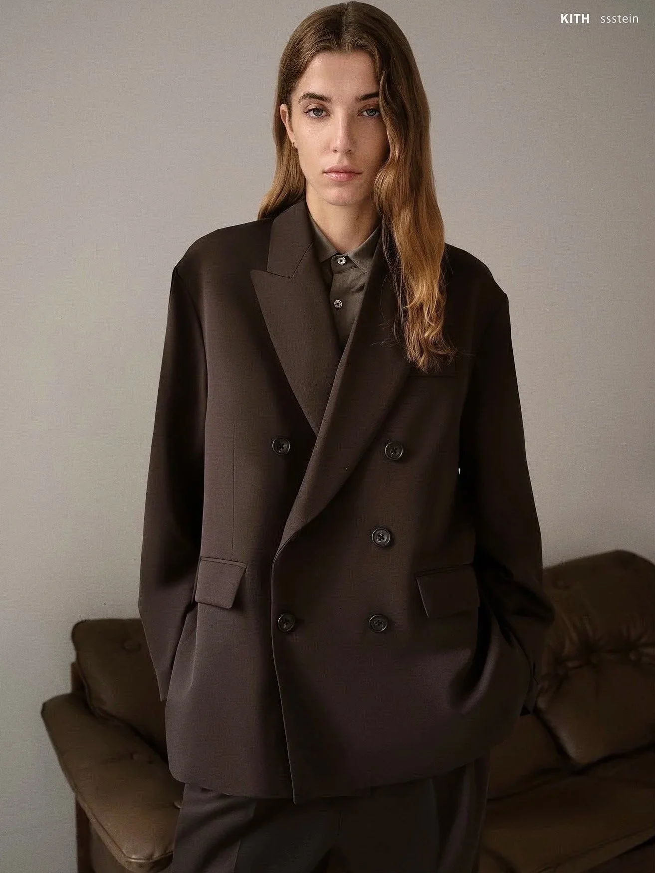 A woman with long, wavy hair, wearing a dark brown oversized double-breasted blazer over a shiny gray shirt.