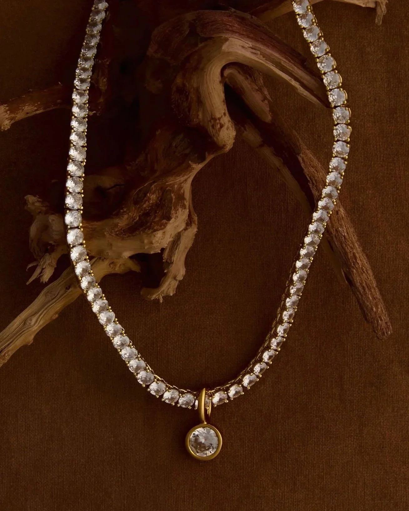Diamond necklace with a large central gemstone pendant resting on a piece of wood, against a brown fabric background.