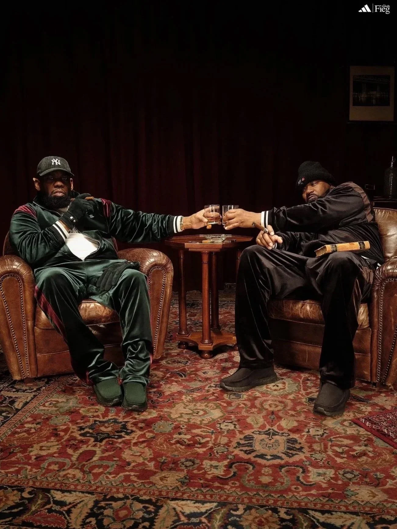 Two men sitting in leather armchairs, toasting with glasses, in a dimly lit room with a patterned rug, a small wooden table with cigars, and a dark red curtain in the background. One man wears a New York Yankees cap and black satin tracksuit, the oth