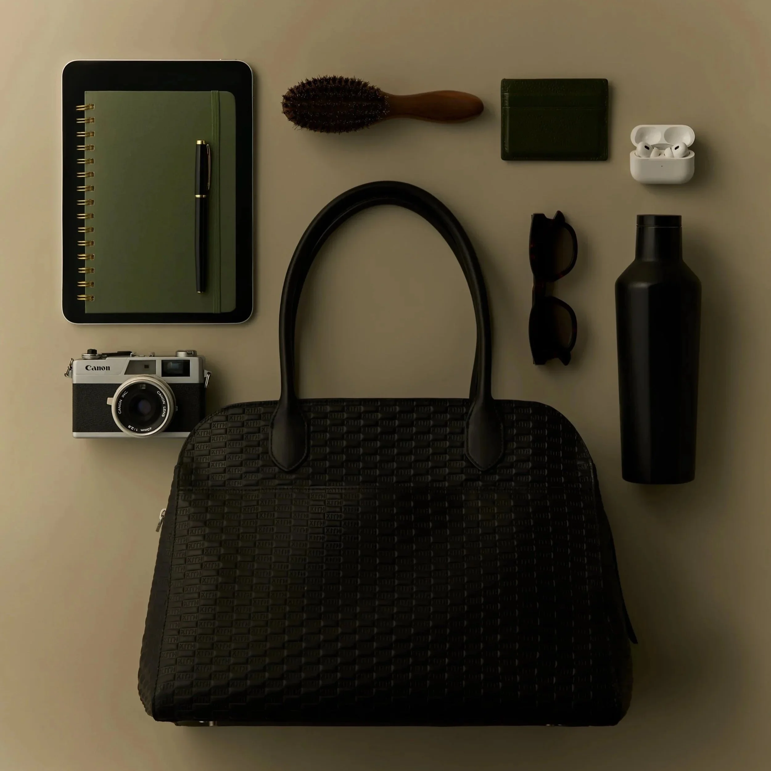 Flat lay of various items including a green notebook with a pen, hairbrush, green cardholder, wireless earbuds in case, sunglasses, black water bottle, and a black textured bag on a light-colored surface.