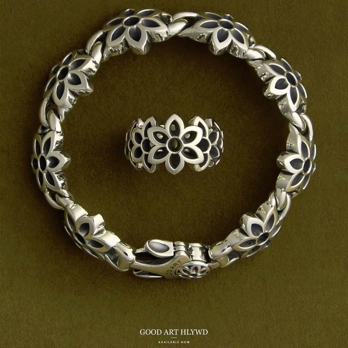 Silver jewelry set including a necklace, ring, and bracelet, all with floral cut-out designs, displayed on a dark background with the text 'GOOD ART HLYWD AVAILABLE NOW' at the bottom.