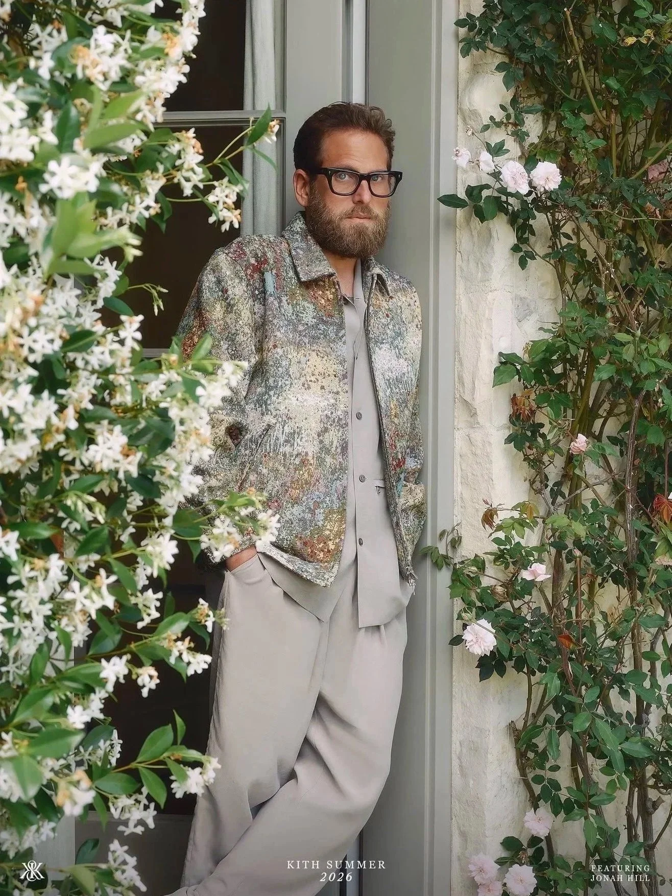 A man with glasses and a beard stands in a doorway surrounded by flowering plants and greenery. He is wearing a multicolored textured jacket and beige pants, looking at the camera with a serious expression.