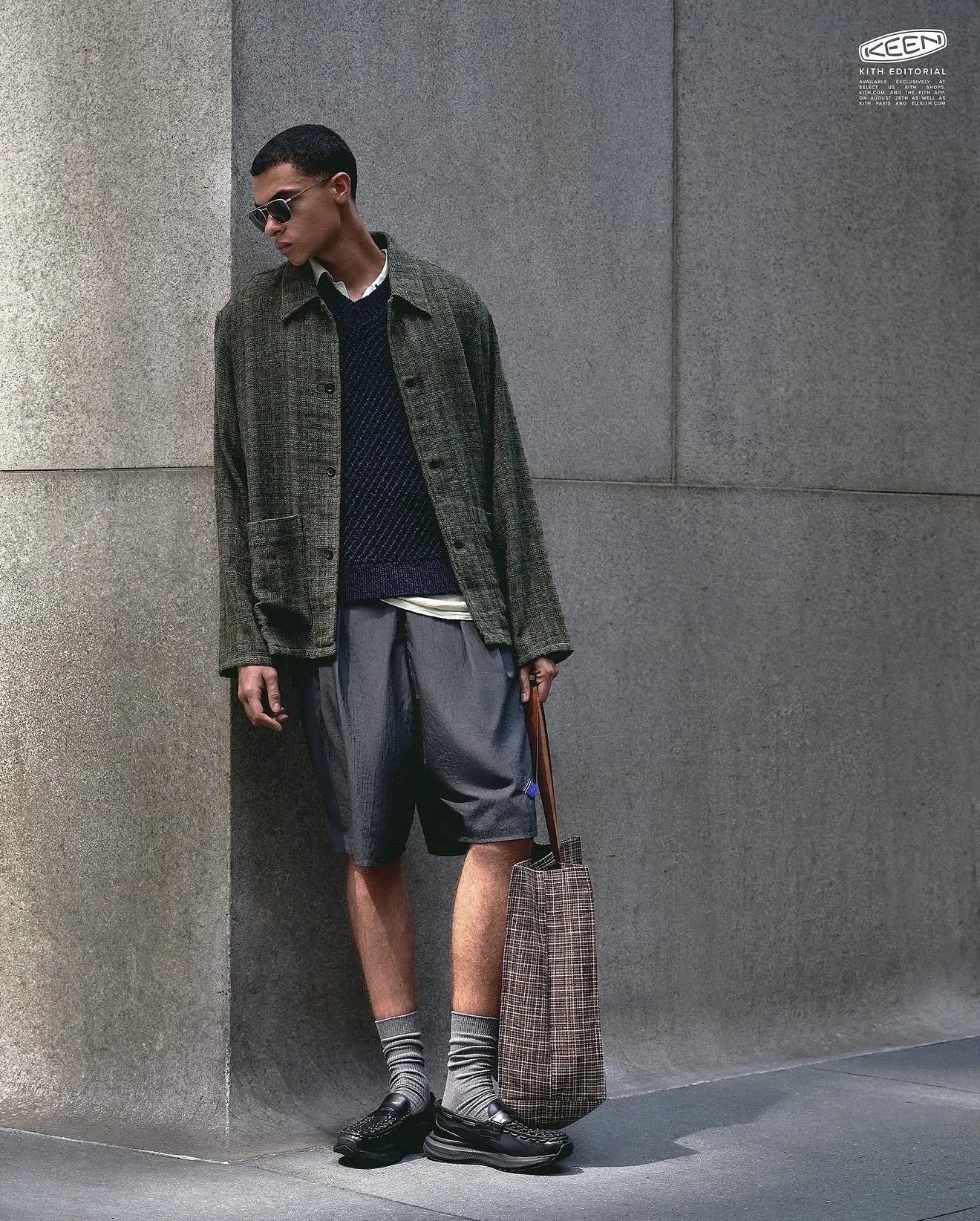 Young man standing against a gray wall, wearing sunglasses, a plaid jacket over a sweater and collared shirt, shorts, striped socks, and loafers, holding a checkered shopping bag.