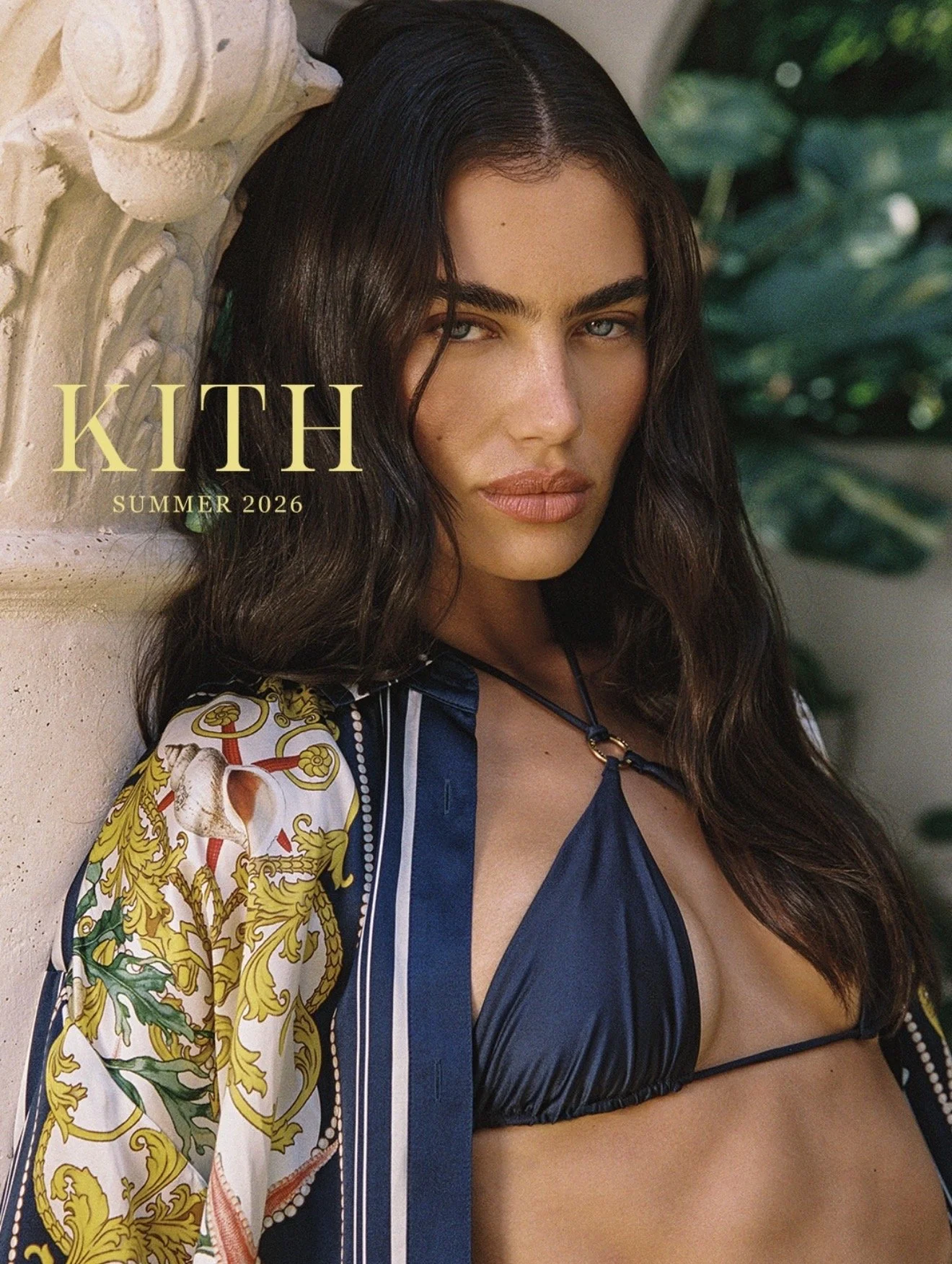 A woman with long dark hair and blue eyes leaning against a beige stone column, wearing a dark blue bikini top and a colorful printed jacket with gold and green designs, with foliage in the background. Text reads "KITH SUMMER 2026".