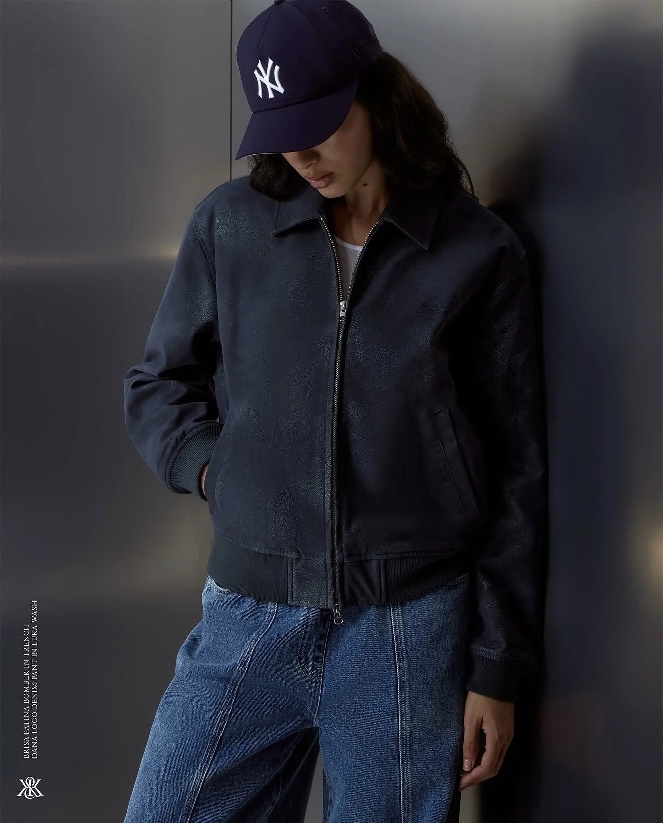 Woman wearing a navy blue New York Yankees cap, black leather jacket, and blue jeans, standing against a gray metallic wall with her hands in her pockets.