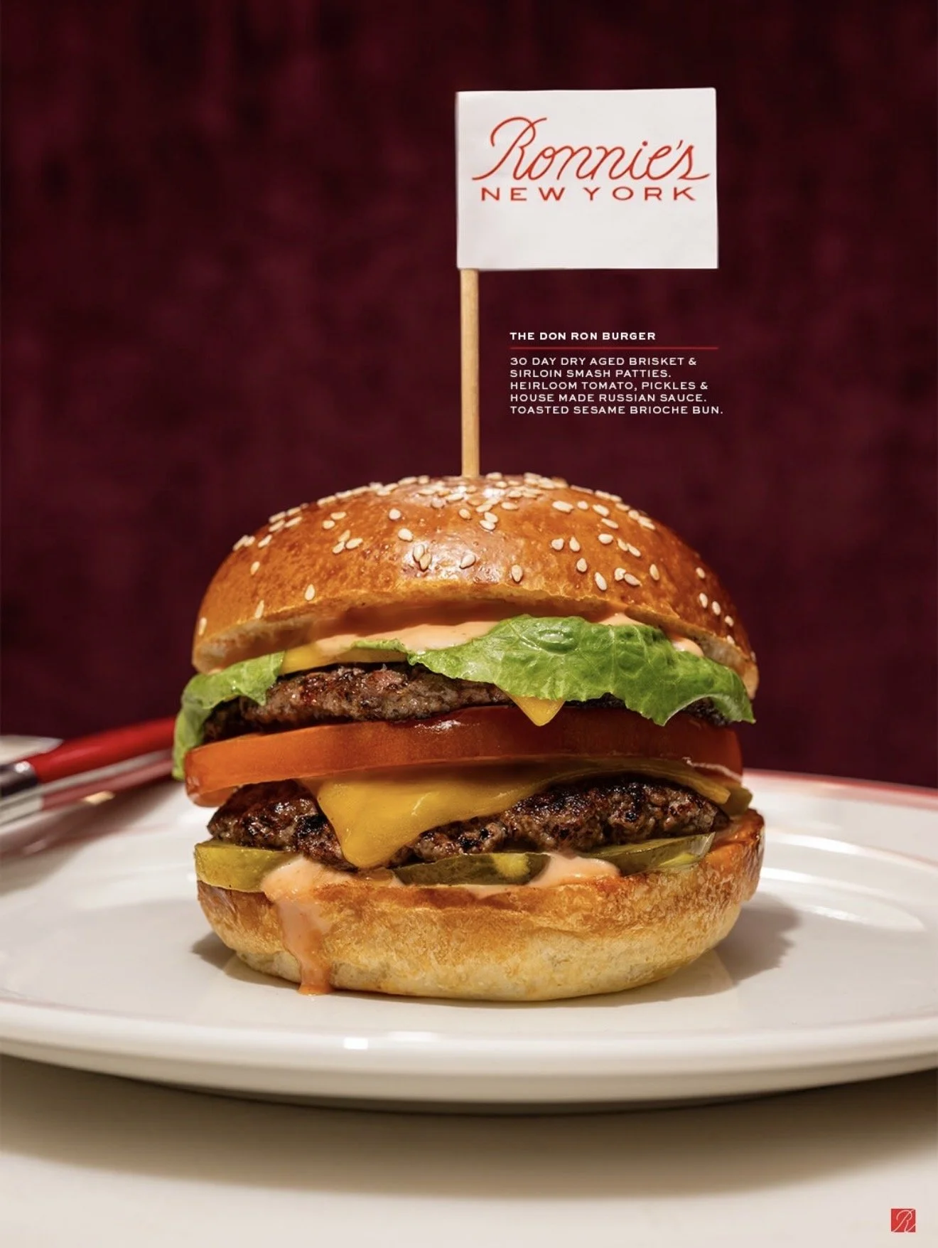 A large cheeseburger with various fresh vegetables layered inside a sesame seed bun, served on a white plate.