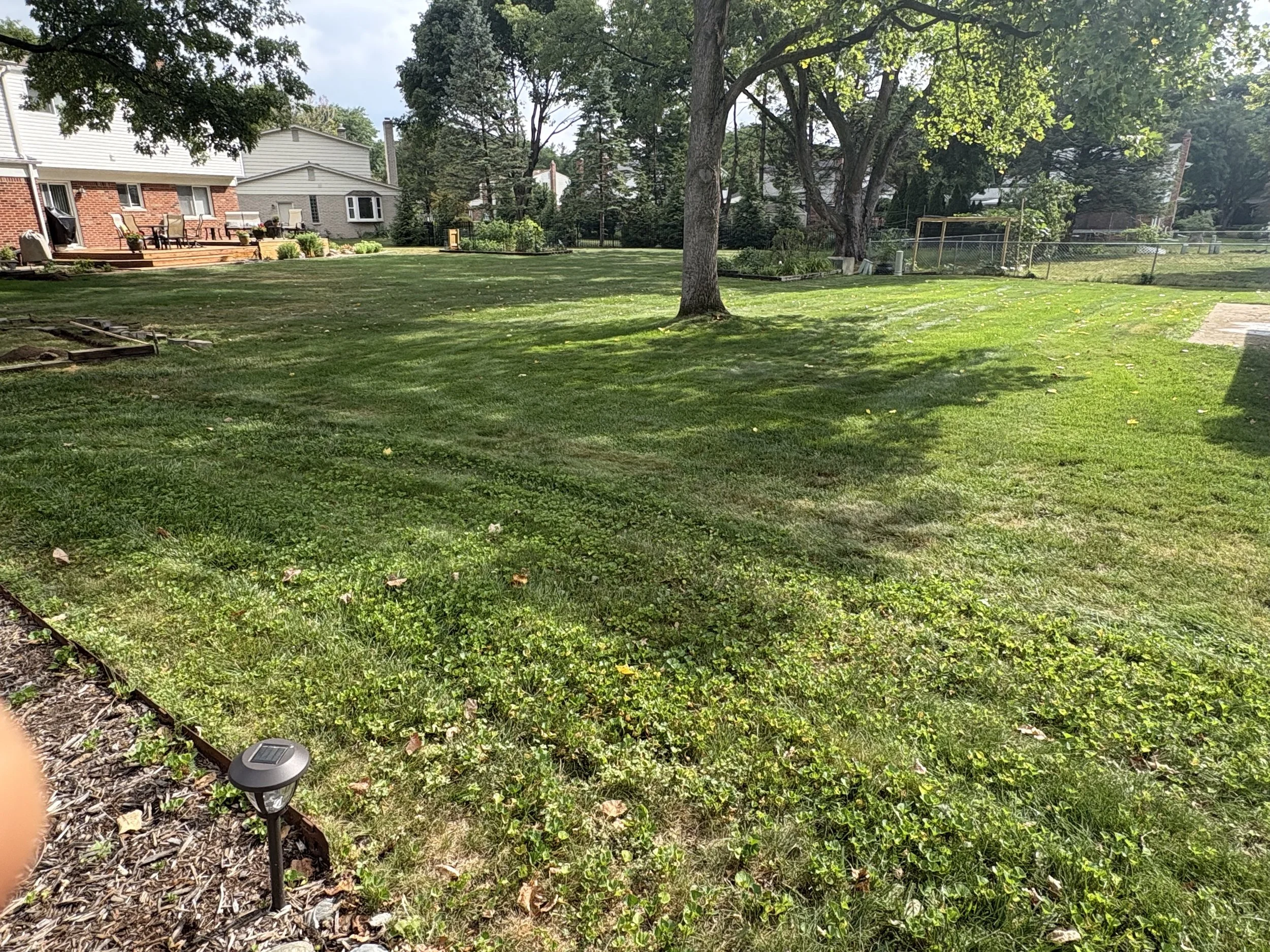 A backyard with a grassy lawn, large trees providing shade, a brick house with a deck and outdoor seating, a fenced garden, and solar path lights along the garden border.