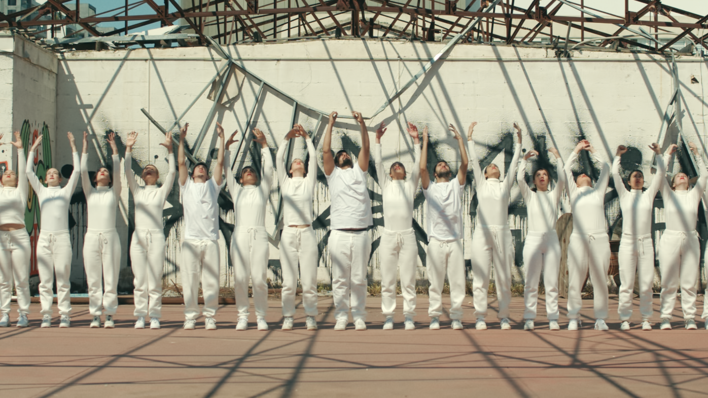 A group of people dressed in white standing outdoors, raising their arms in the air, with a damaged structure and graffiti on the wall behind them.