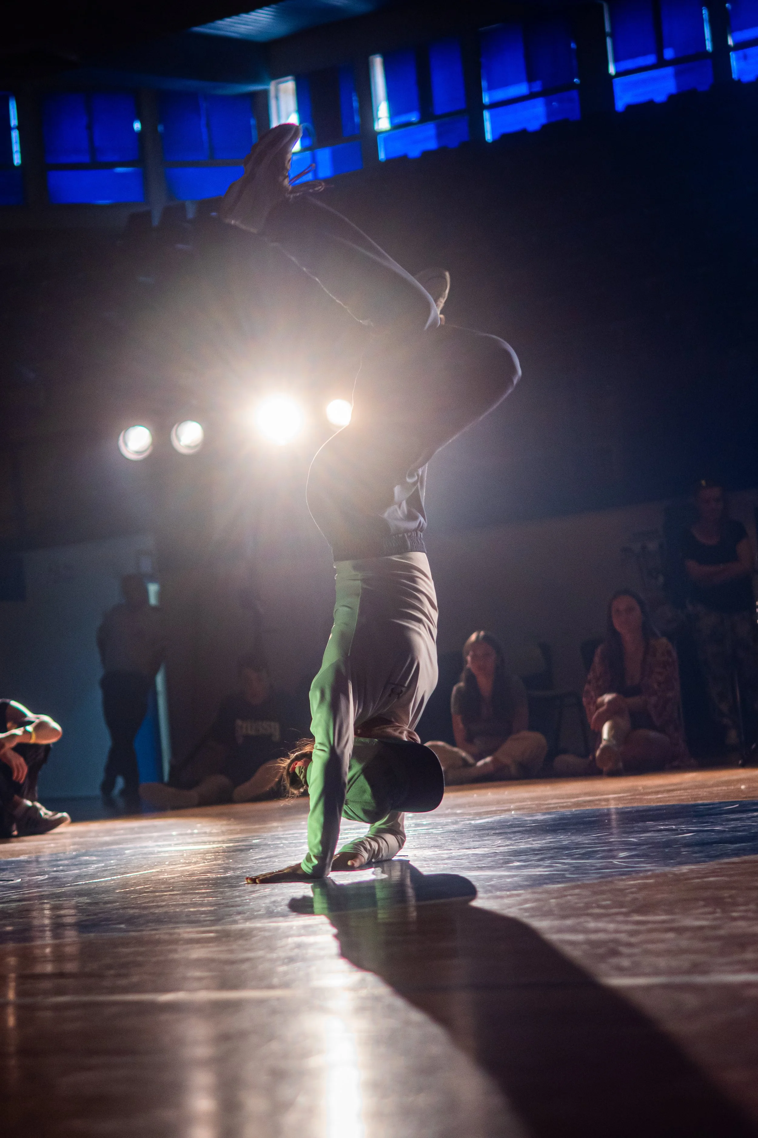 A dancer performing a handstand on a stage with an audience watching. The background is dark with blue lights through windows, and the stage is lit by bright lights.