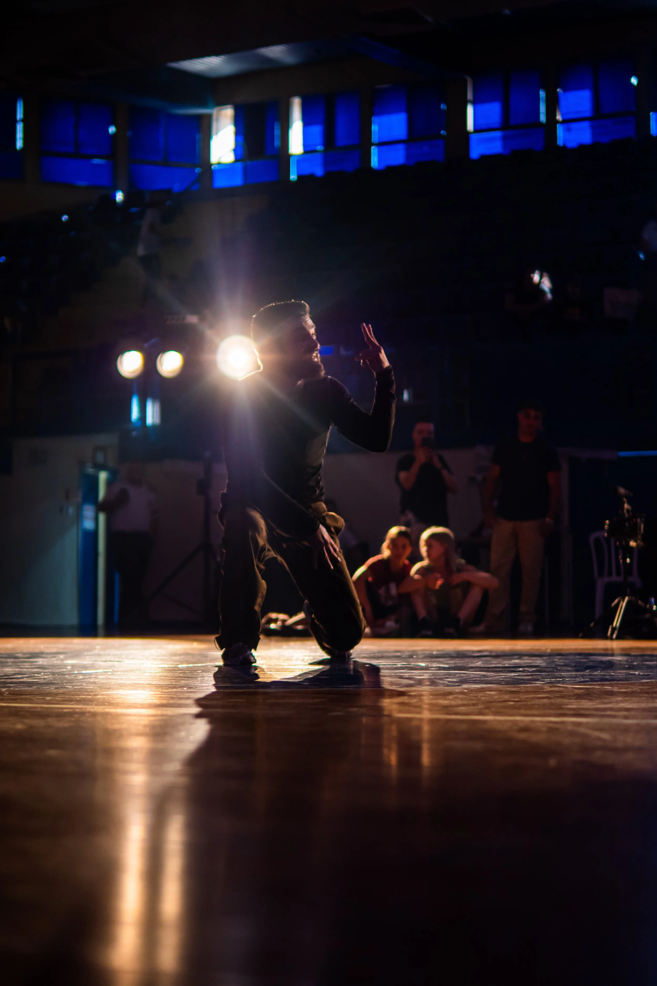 A male dancer performing on a dimly lit stage with bright lighting behind him, surrounded by an audience.