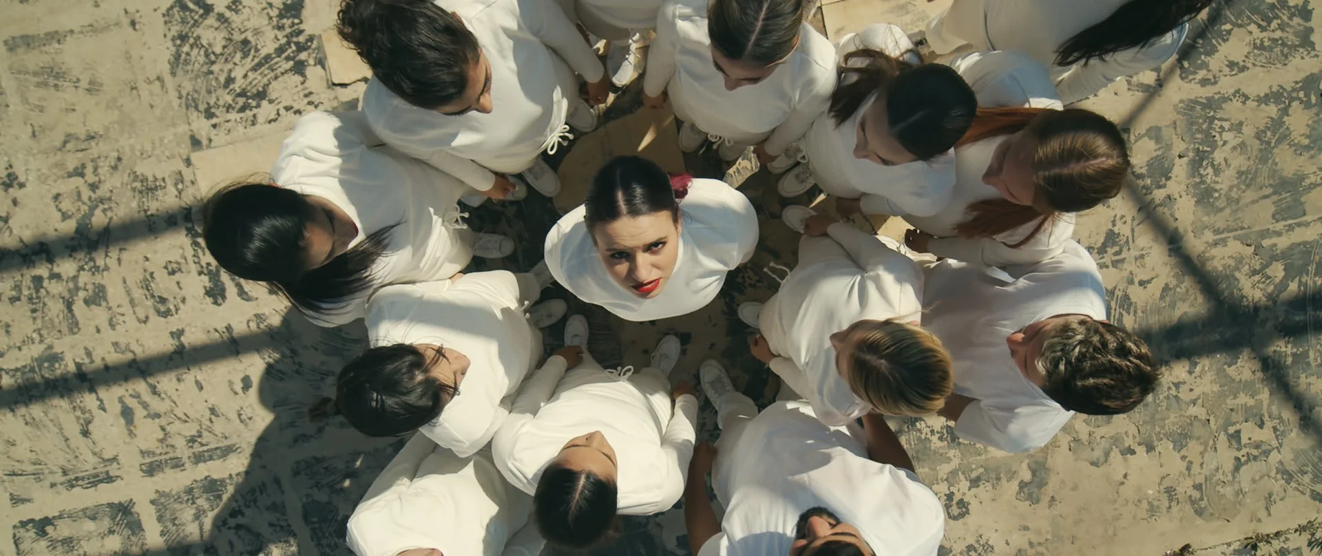 A group of women dressed in white among chain-link fence shadows, looking up at the camera with one woman at the center looking directly.