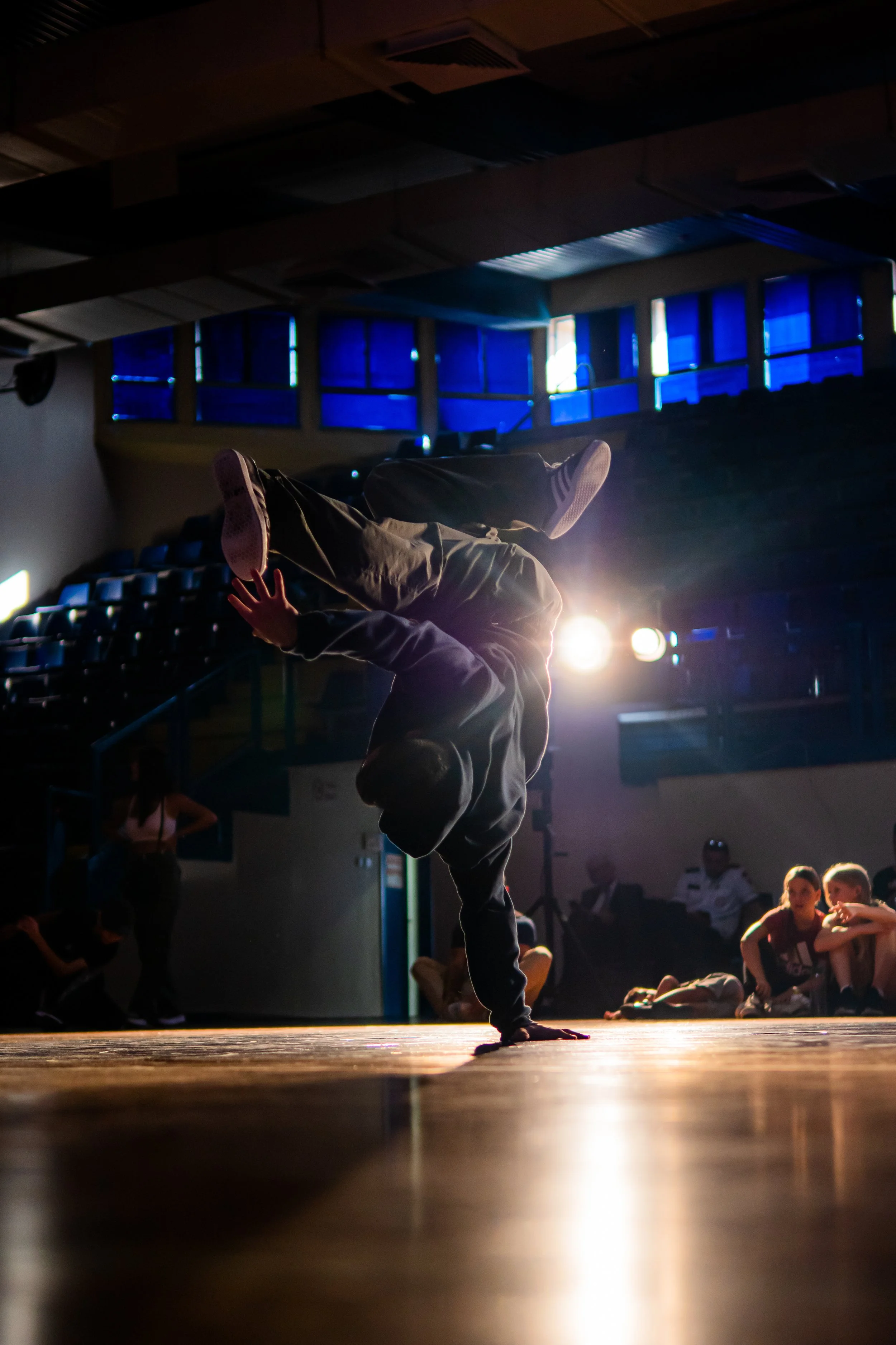 A person performing a breakdancing move on stage, with a group of people sitting and standing in the background. The scene is lit with bright stage lights, and blue windows are visible in the background.