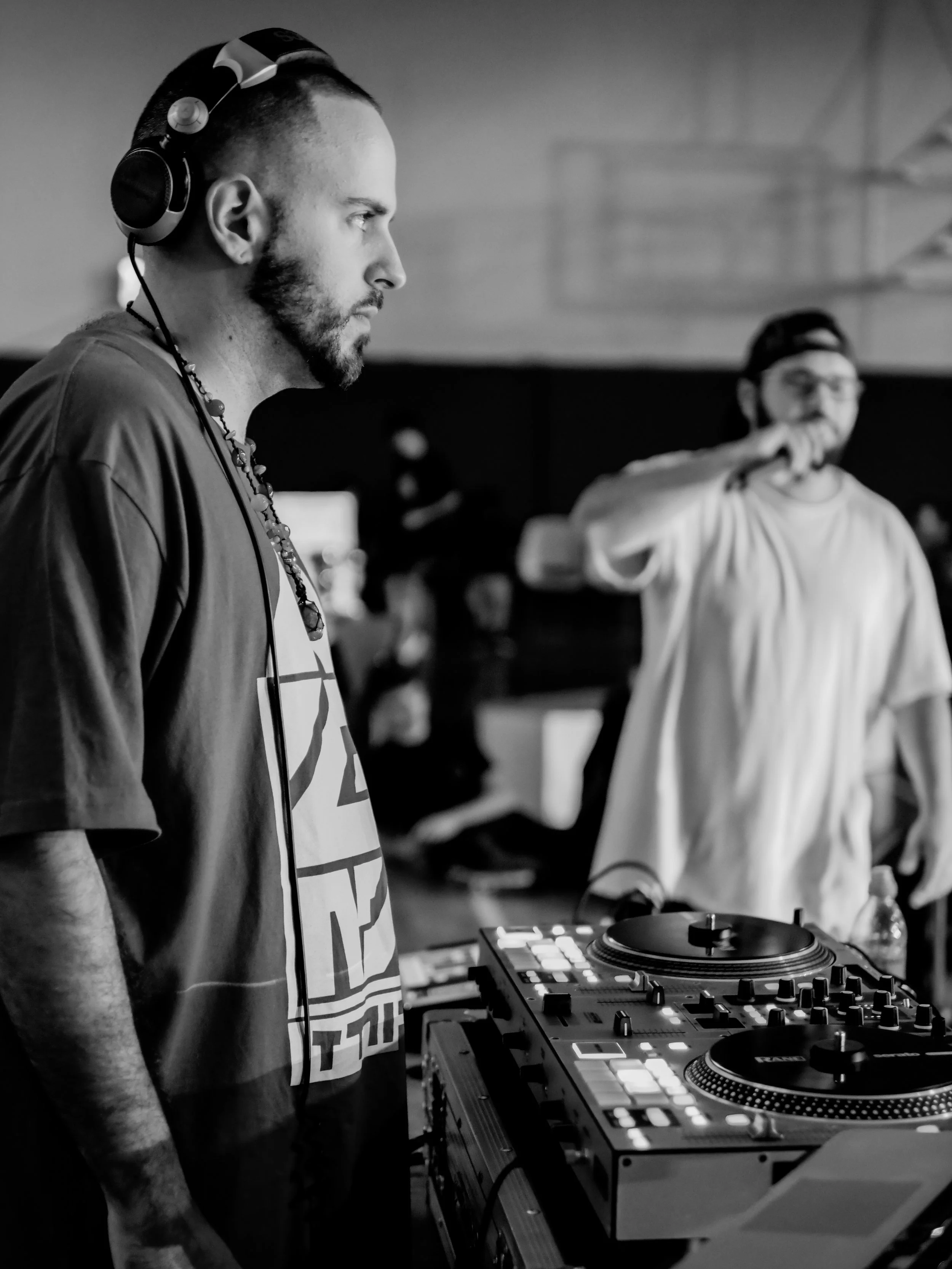 A DJ with a beard wearing headphones and a graphic t-shirt, standing at a DJ mixer in a dimly lit room, with another person wearing glasses and a cap in the background.