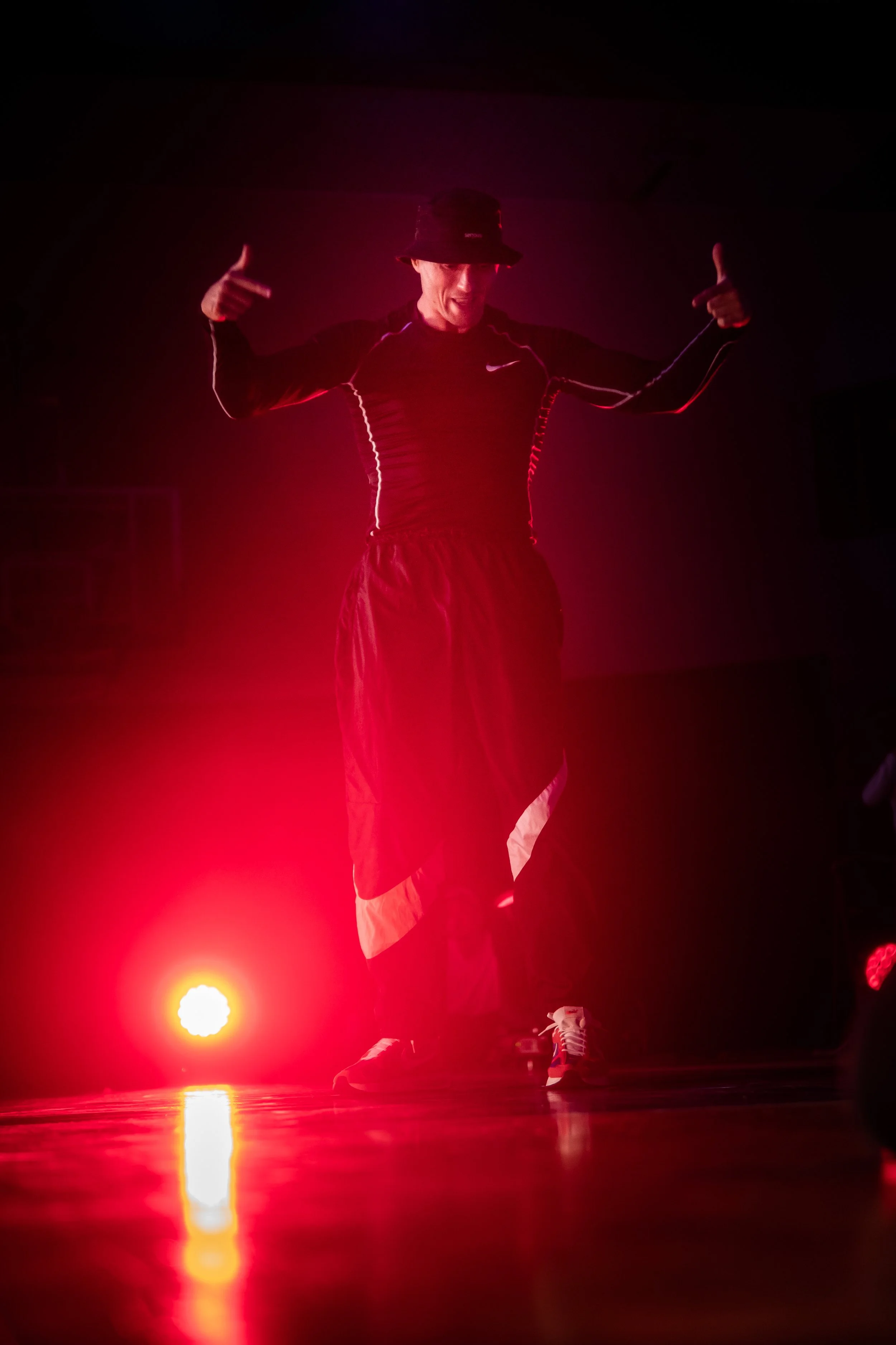 A man wearing a black bucket hat, black Nike long sleeve shirt, and light-colored shorts, standing on a dark stage illuminated by red stage lights. He is smiling, pointing to himself with both hands, and wearing colorful sneakers. The stage has a reflective surface with a bright red light and a yellow spotlight visible.